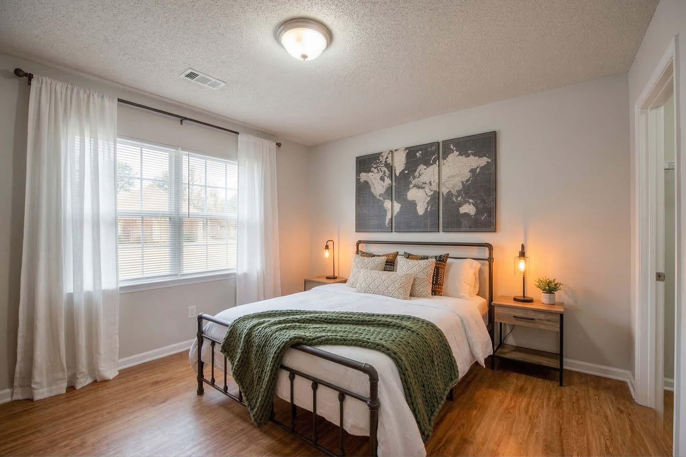 A cozy bedroom featuring a large bed with white linens and a green throw, two bedside tables with lamps, a world map on the wall, and a window with sheer curtains. The room has wooden flooring and neutral-colored walls, giving it a warm and inviting atmosphere.