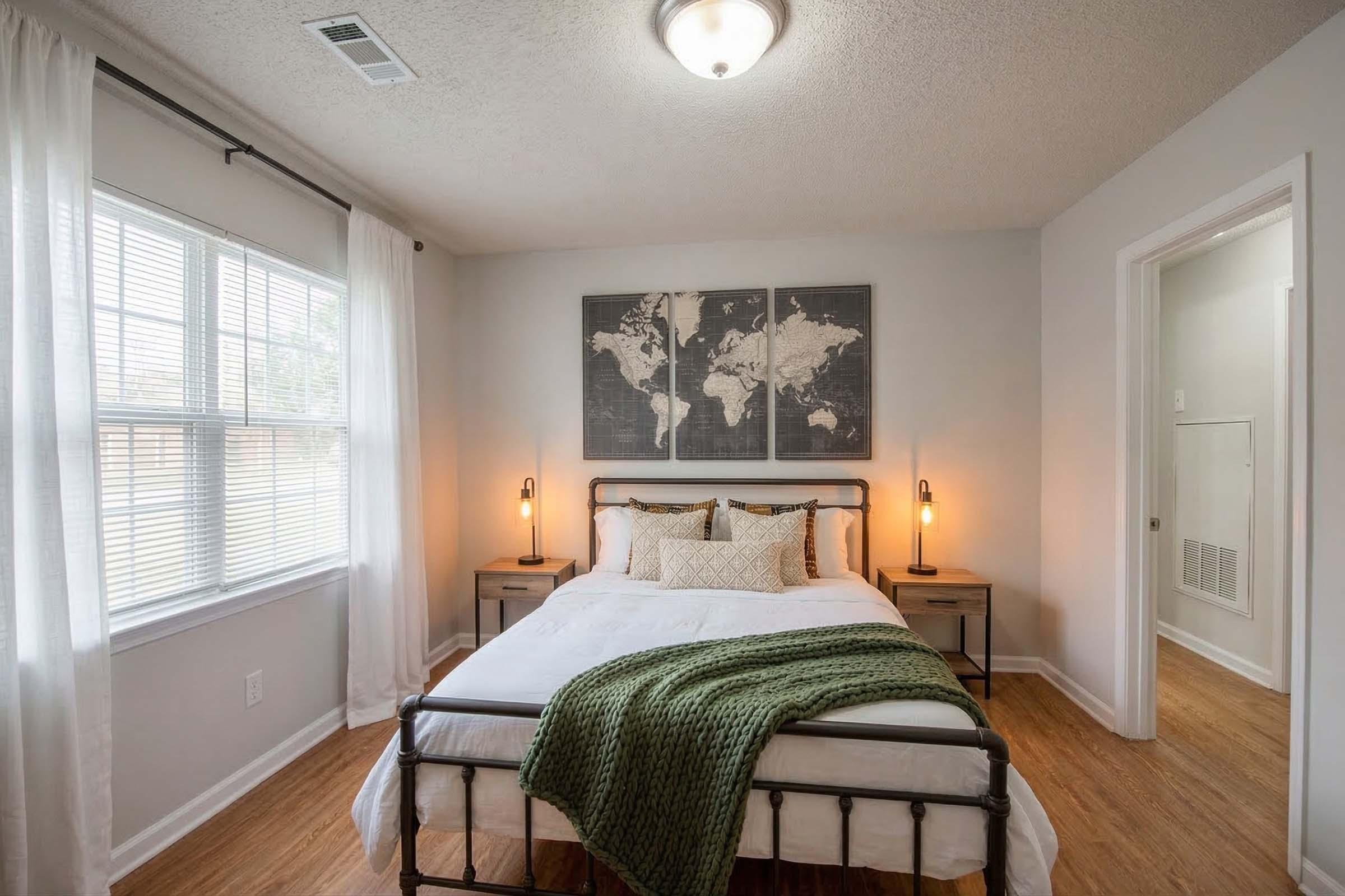 A cozy bedroom featuring a double bed with a green throw blanket, flanked by two stylish bedside tables with lamps. A large world map art piece hangs on the wall, and the room has large windows allowing natural light to brighten the space. The flooring is wooden, adding warmth to the ambiance.