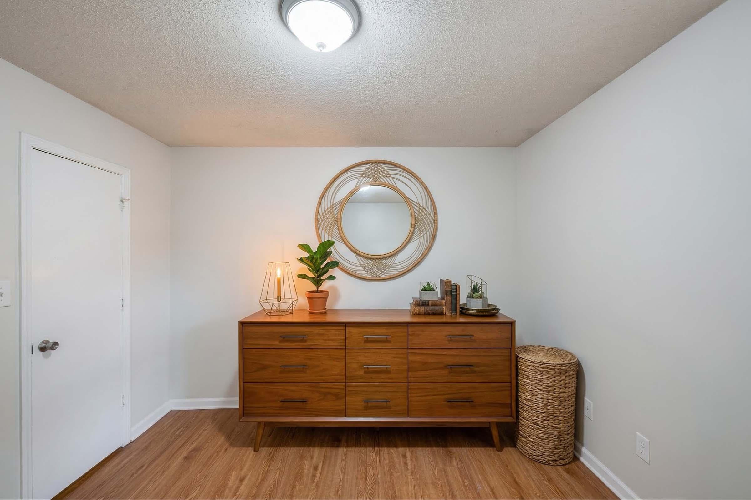 A modern interior scene featuring a stylish wooden dresser with multiple drawers, topped with decorative items like a potted plant and a small tray. A round mirror with a woven frame hangs above the dresser, and a soft light fixture illuminates the space. A woven basket is placed nearby, adding texture to the room.