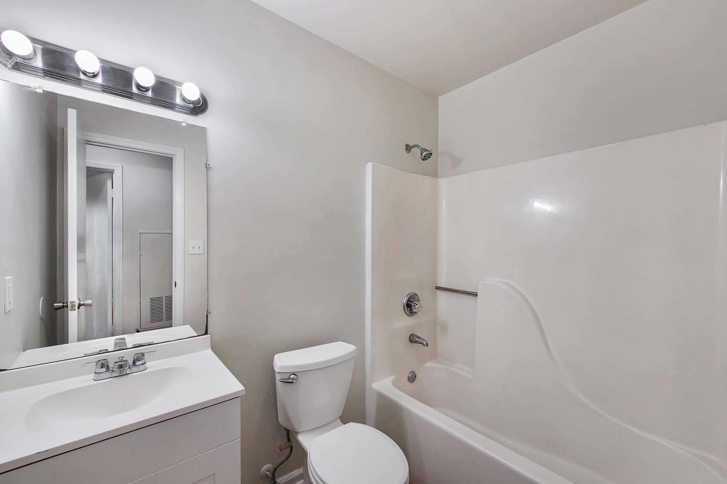 A clean, modern bathroom featuring a white bathtub and shower combination, a toilet, and a sink with a mirror above. The lighting consists of five bulbs mounted above the mirror. The walls are a neutral color, and there is a door leading to another room visible in the background.
