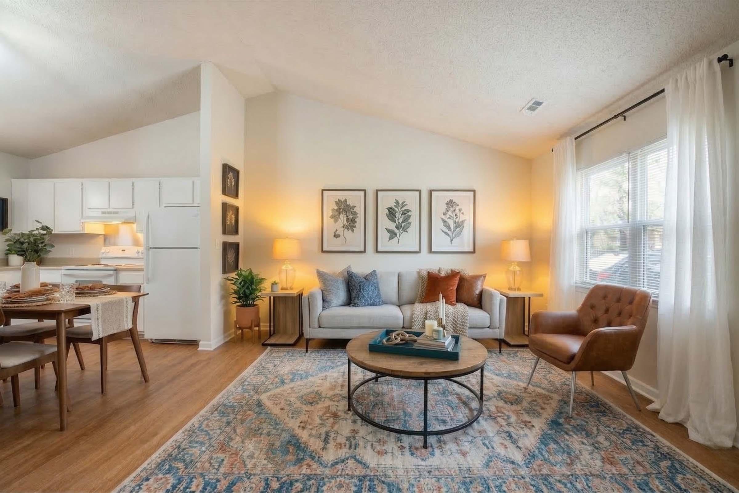 A well-decorated living room featuring a light-colored couch with decorative pillows, a round coffee table with a decorative centerpiece, a cozy armchair, and a dining area with a wooden table. The walls are adorned with botanical prints, and large windows allow natural light to fill the space.