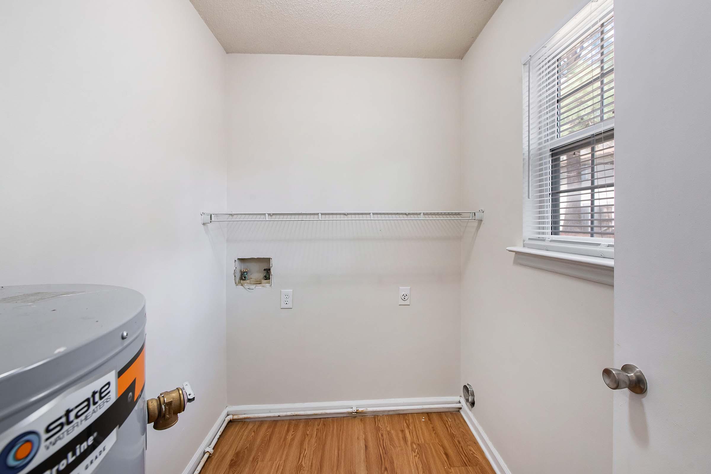 A small, empty laundry room featuring a water heater on the left, a wall with a small hole, and a shelf mounted on the wall. The room has light-colored walls, a window with blinds, and hardwood flooring. There are electrical outlets visible on the wall.