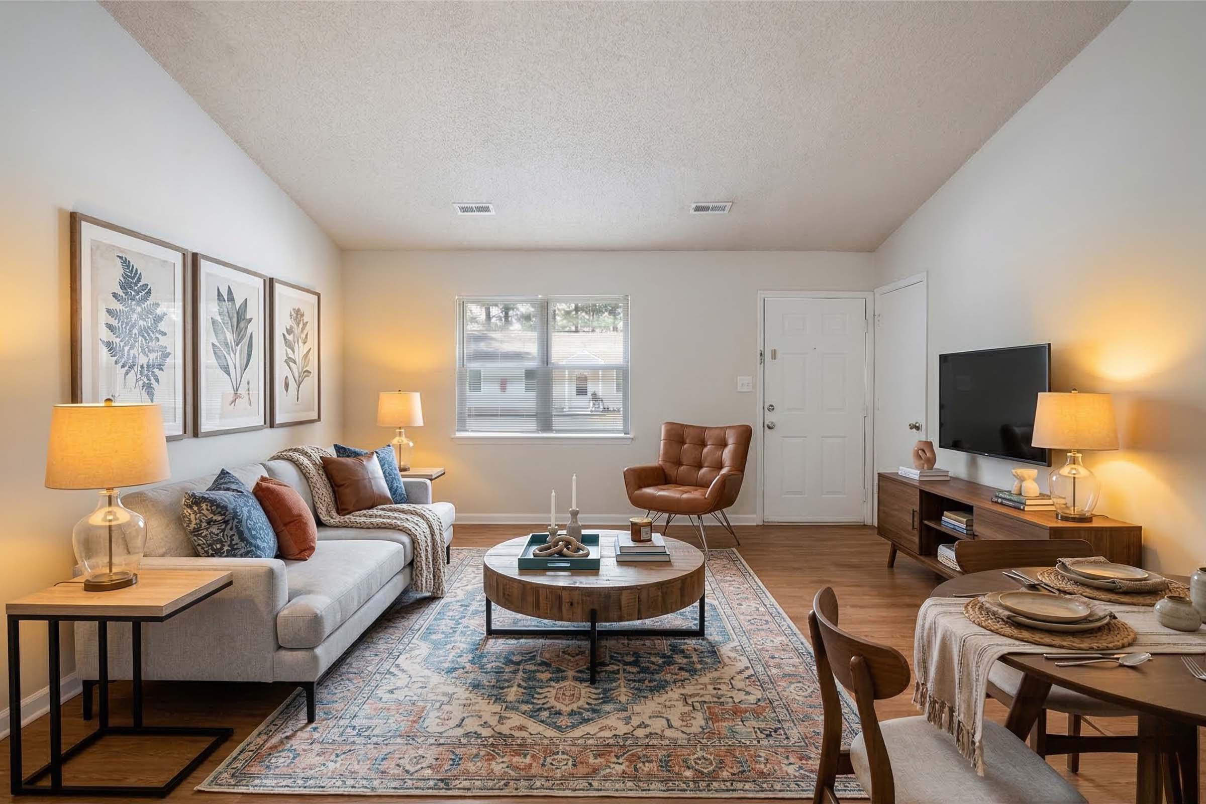 A cozy living room featuring a light gray sofa, an orange armchair, and a wooden coffee table with decorative items. The walls are adorned with framed botanical artwork. A television is mounted on the wall, and there are two lamps providing warm lighting. A dining area with a table and chairs is also visible.