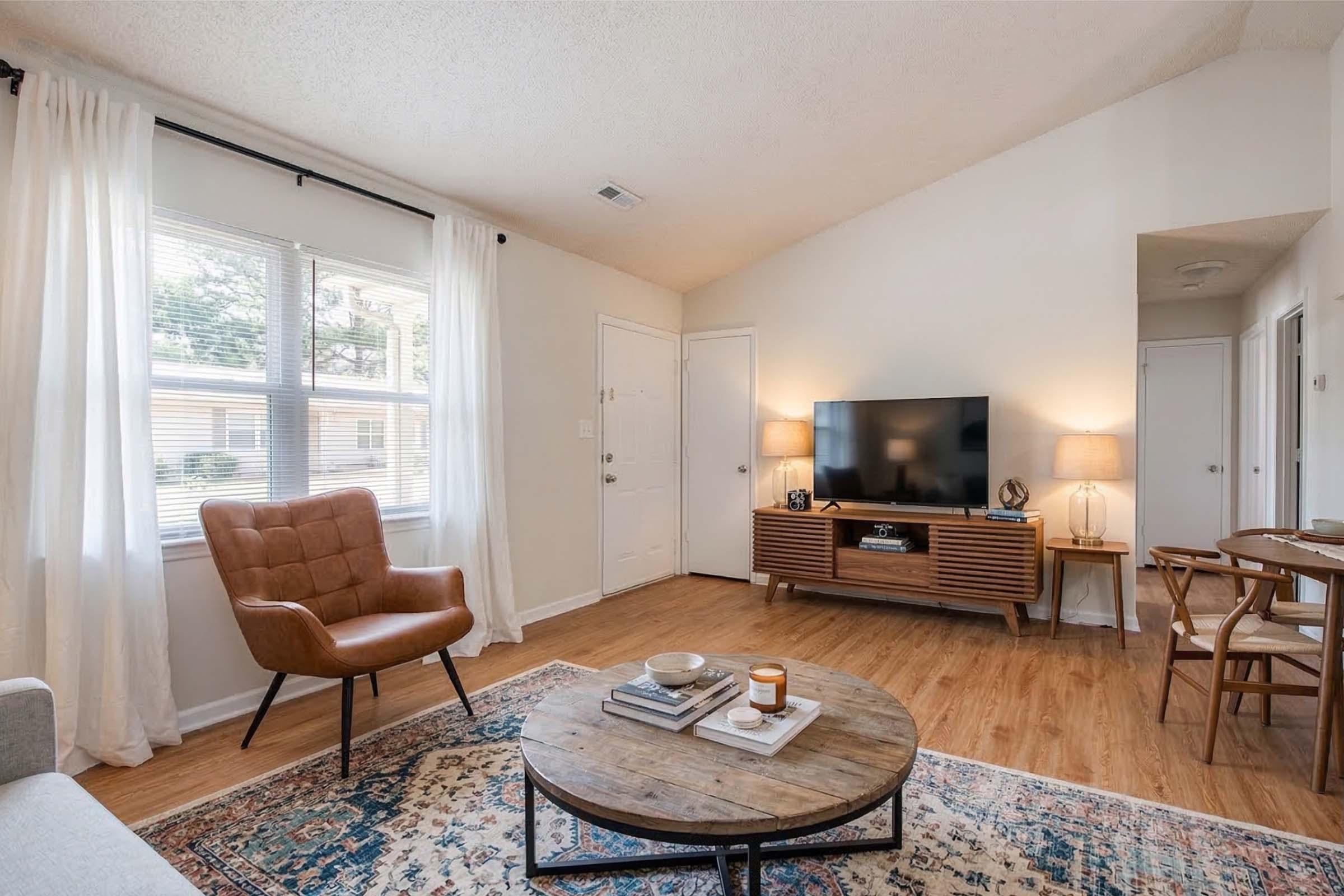 A cozy living room featuring a tan armchair, a round coffee table with decor, a modern TV stand, and a flat-screen television. Large windows let in natural light, with sheer white curtains. The floor is hardwood, and the walls are painted a soft neutral color, creating a warm and inviting atmosphere.