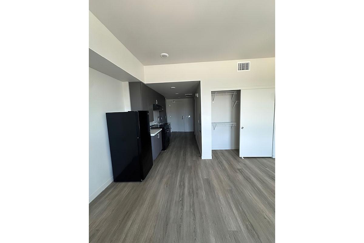 Interior view of a modern apartment with an open layout. The image shows a kitchen area to the left with a black refrigerator, a counter, and cabinets.