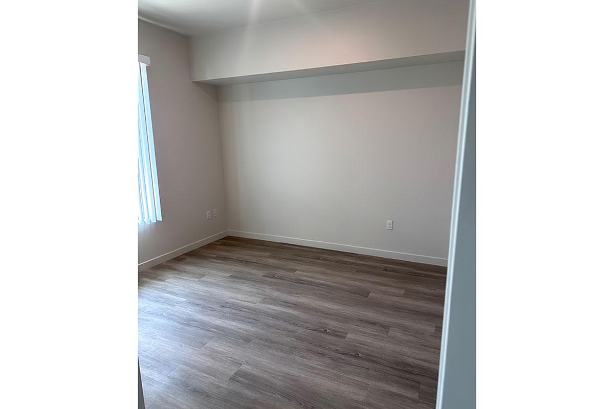 Empty room with light-colored walls and wooden flooring. A window with vertical blinds allows natural light in from the left side.