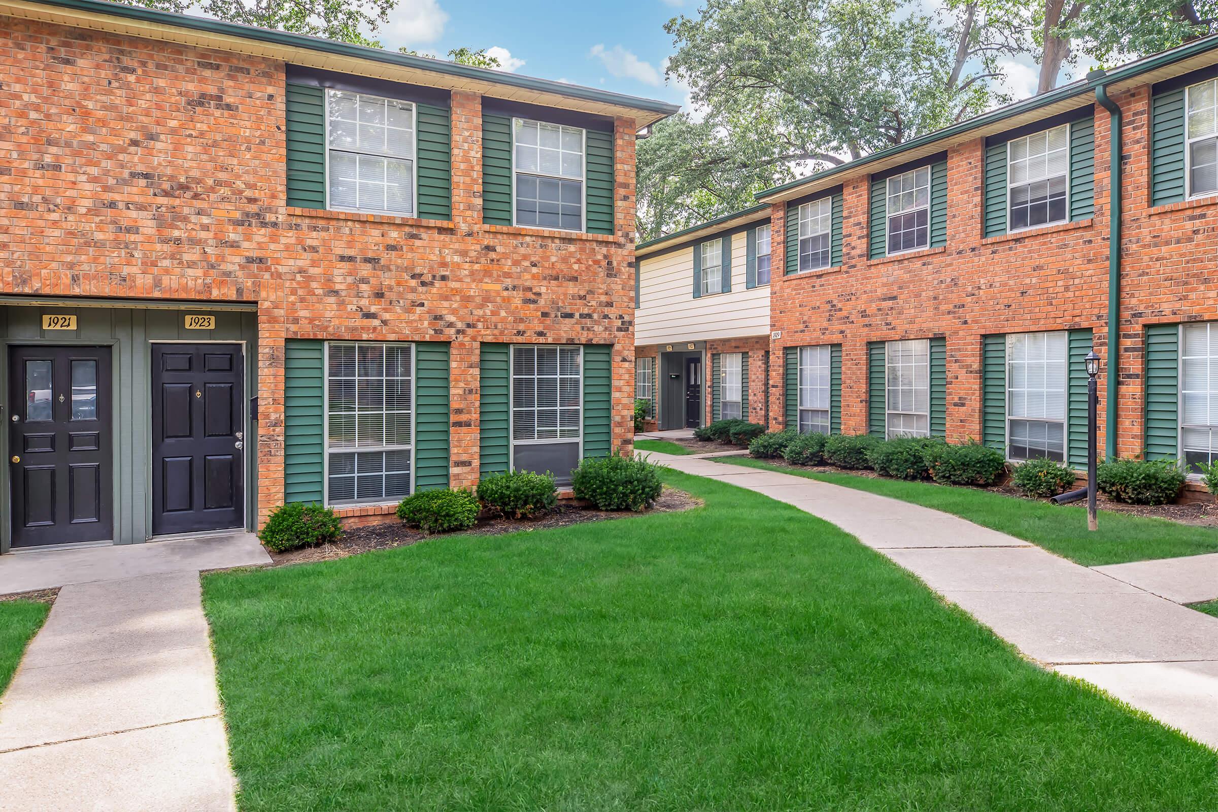 A well-maintained outdoor area with two brick apartment buildings. The foreground features a neatly landscaped lawn with shrubs and a concrete pathway leading to the entrances of the units. The buildings have green shutters and large windows, creating a welcoming atmosphere.