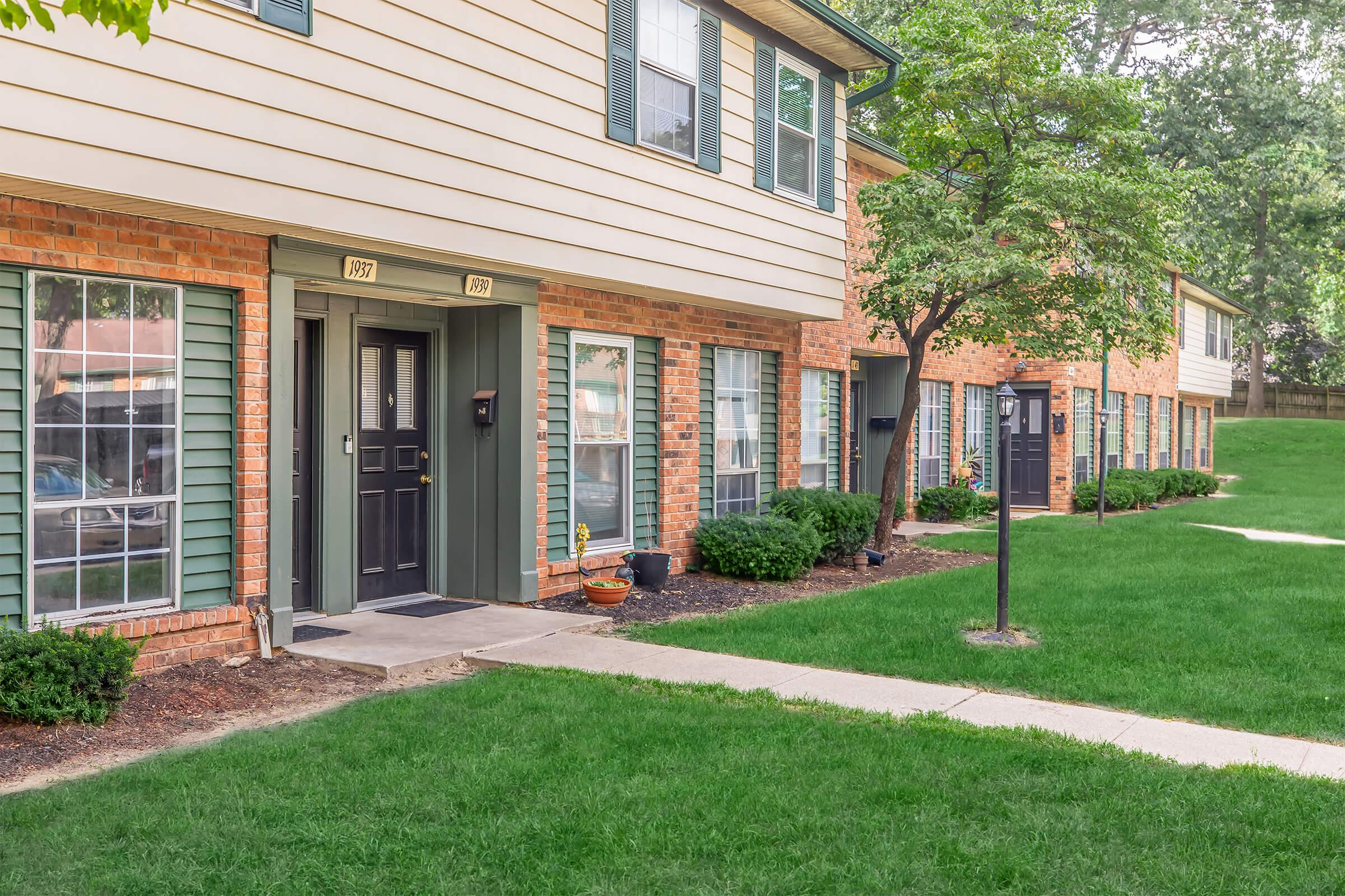 A view of a well-maintained brick apartment complex featuring multiple units with green shutters, grassy lawns, and tree landscaping. The entrance of one unit is highlighted, showcasing a front door with a mailbox. Pathways connect the units, creating a welcoming residential atmosphere.