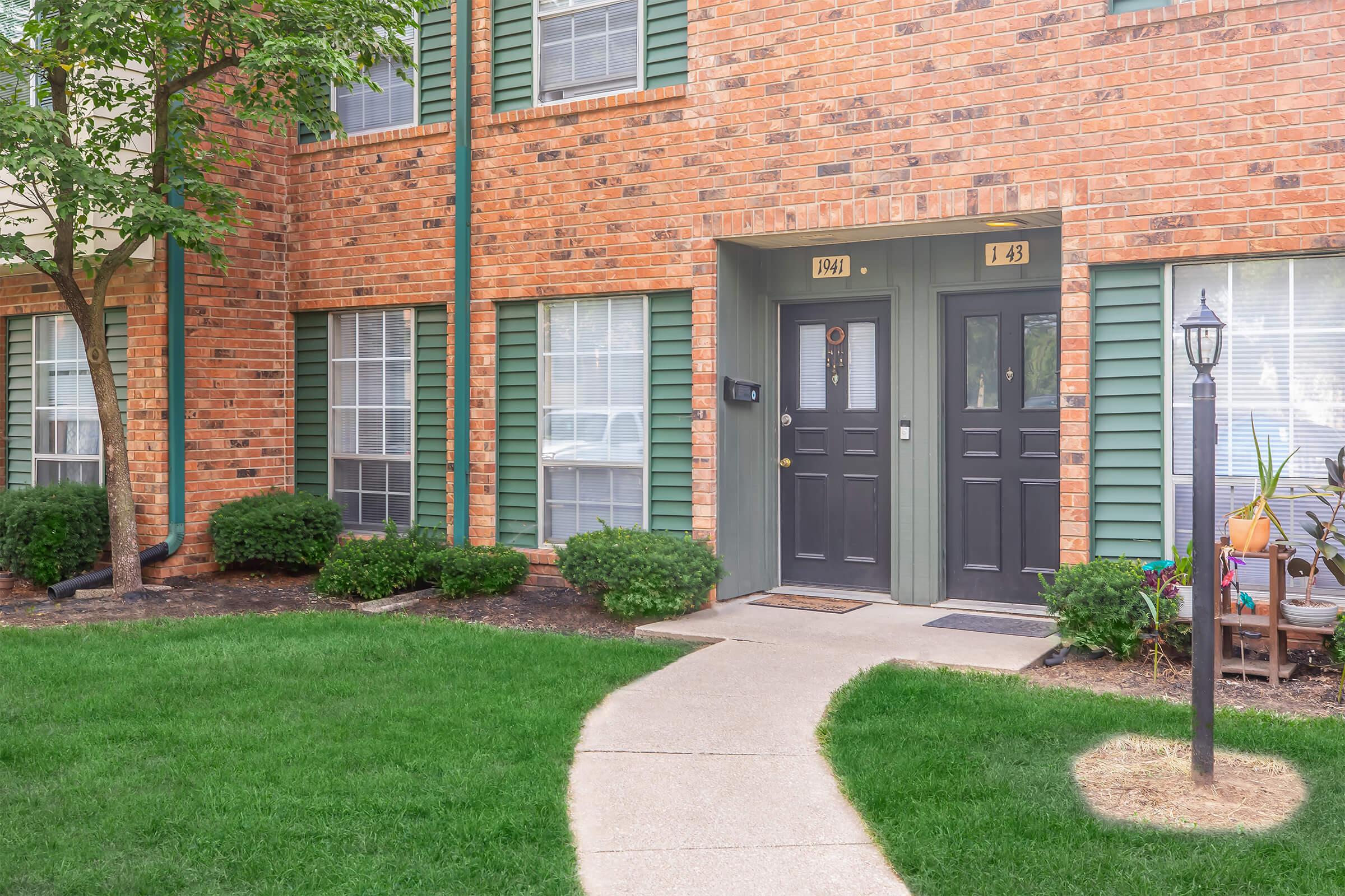 A brick apartment building with two doors, numbered 1943 and 1945, surrounded by green bushes and a pathway leading to the entrance. There's a lamppost near the door on the right, and the windows have green shutters. The lawn is well-maintained.