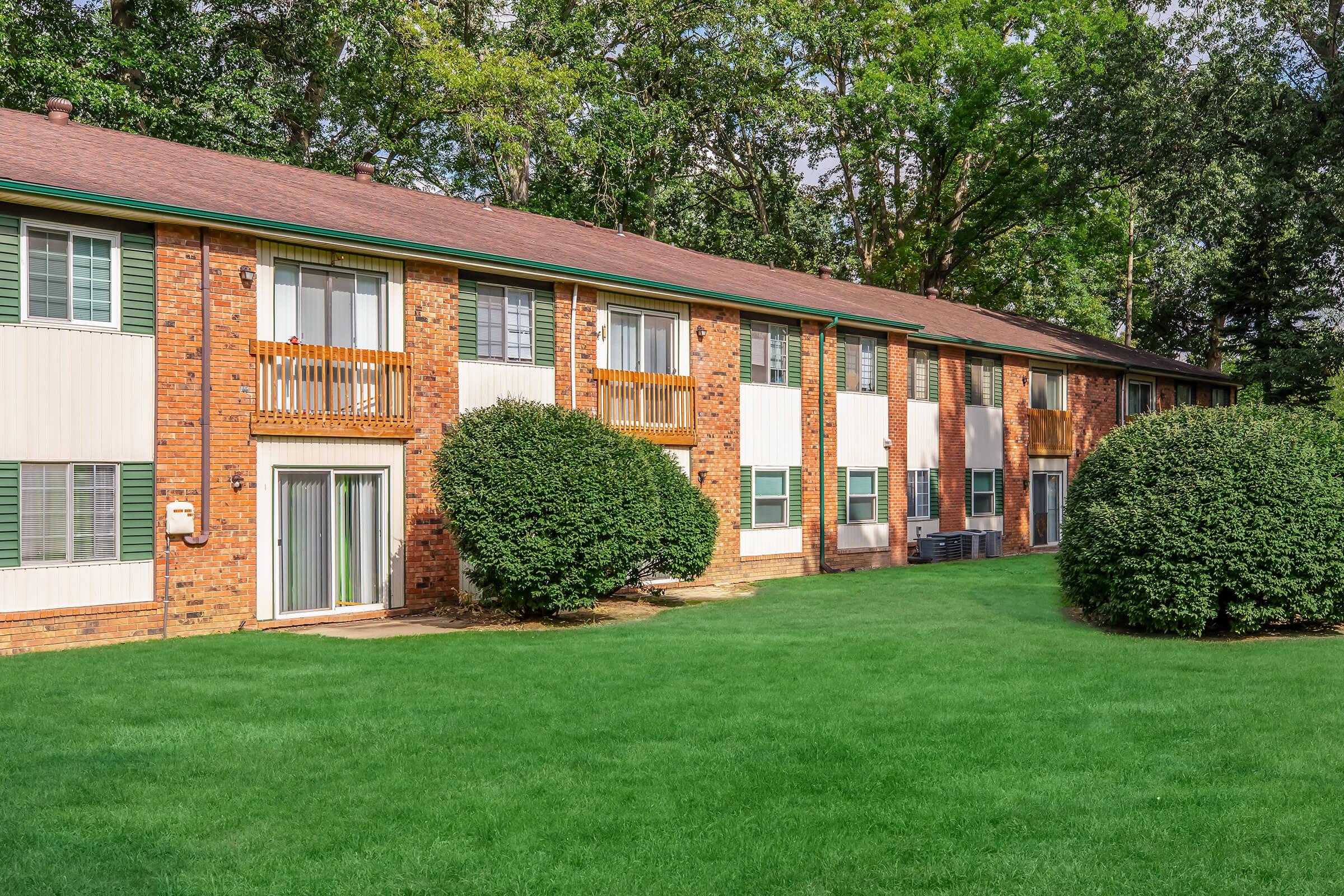 A view of a two-story brick apartment building surrounded by green shrubs and a well-maintained lawn. The building features multiple windows with shutters and balconies, set against a backdrop of trees. The scene is bright and inviting, suggesting a peaceful residential environment.