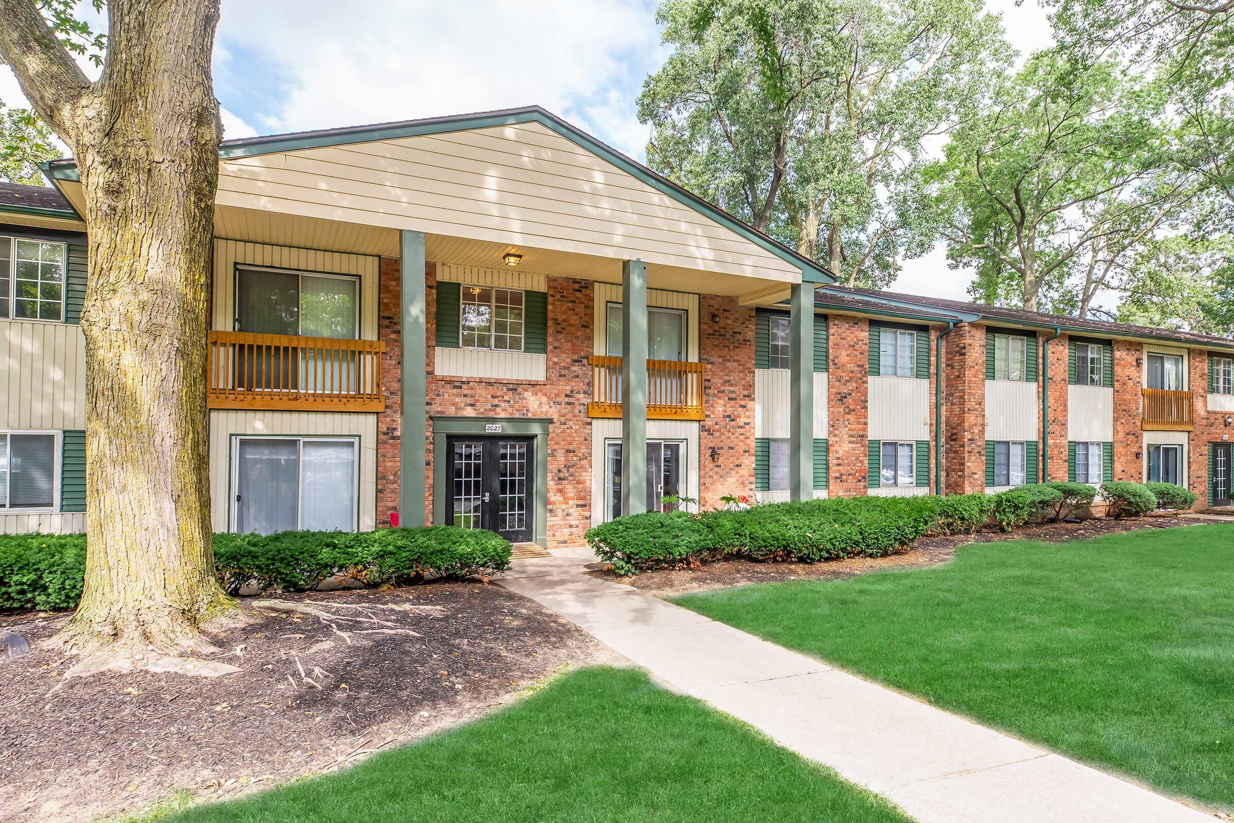 A two-story apartment building featuring a brick and siding facade. The entrance has a covered porch with two balconies above. The landscaping includes well-maintained green grass, small shrubs, and a tree nearby. The sky is partly cloudy, creating a pleasant outdoor atmosphere.