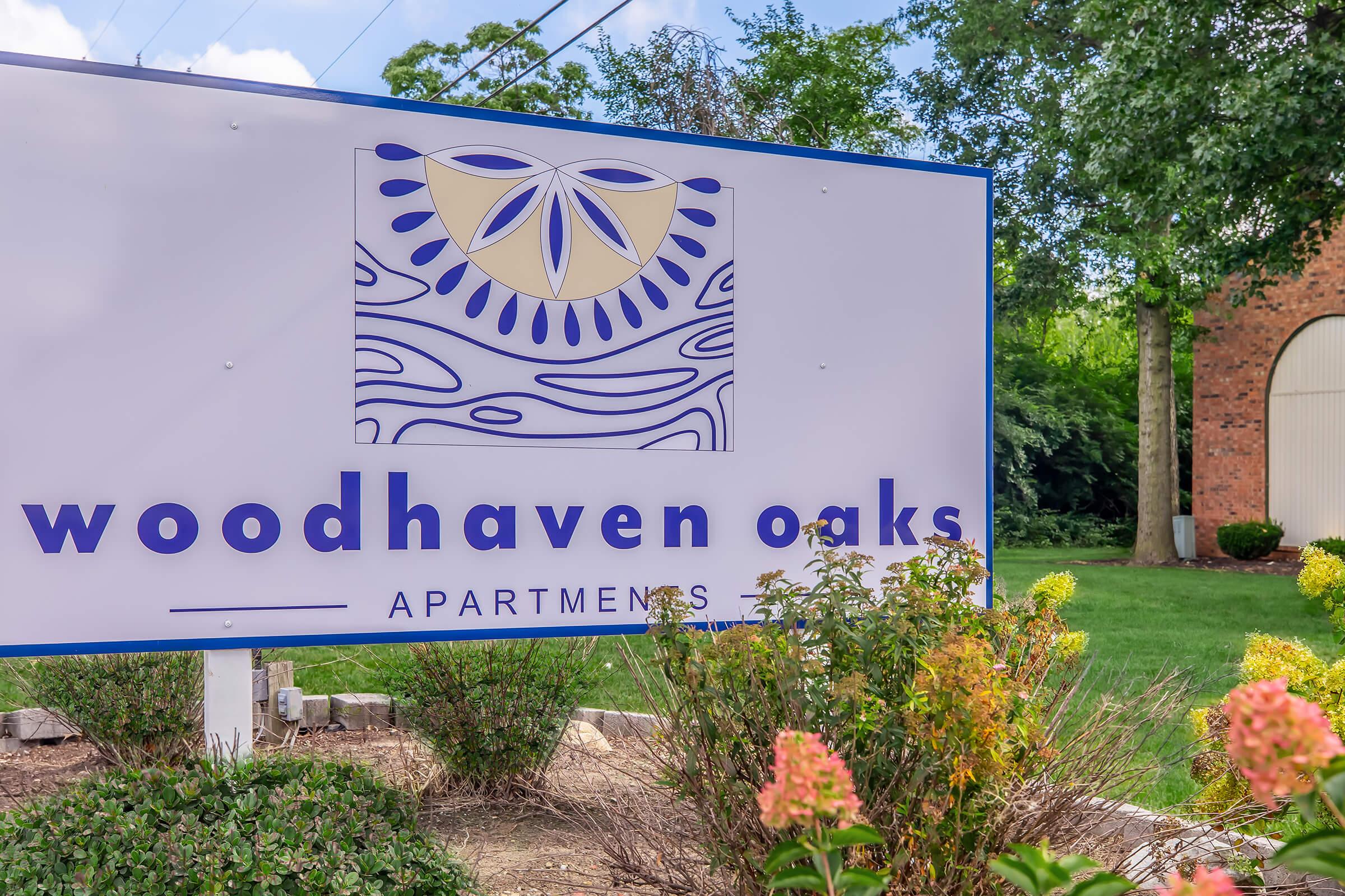 Sign for Woodhaven Oaks Apartments featuring a decorative logo and the text "woodhaven oaks APARTMENTS." The sign is surrounded by green grass and flowering bushes under a blue sky with scattered clouds.