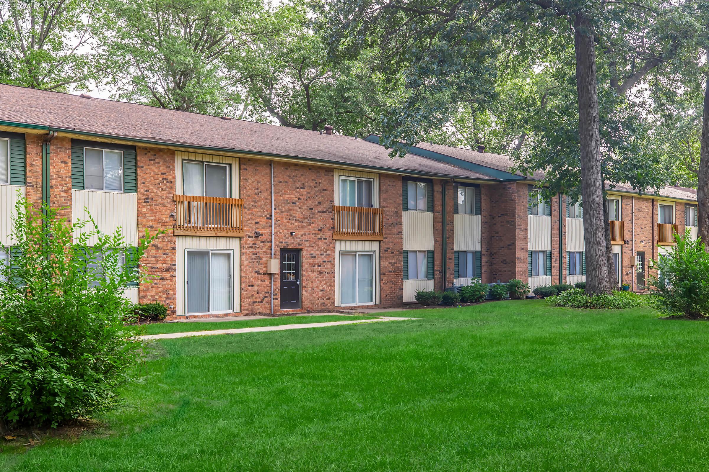 A two-story apartment building with a brick exterior, featuring balconies and large windows. The surrounding area has well-maintained green grass and small bushes. Trees provide shade along the sides, creating a serene residential setting.