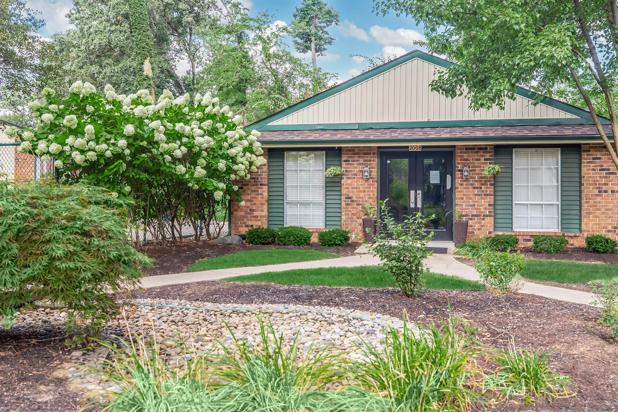 A brick building with a green roof, surrounded by well-maintained landscaping, including flowering shrubs and a stone path leading to the entrance. The windows are covered with white curtains, and the surrounding area has trees and grass, creating a pleasant outdoor setting.