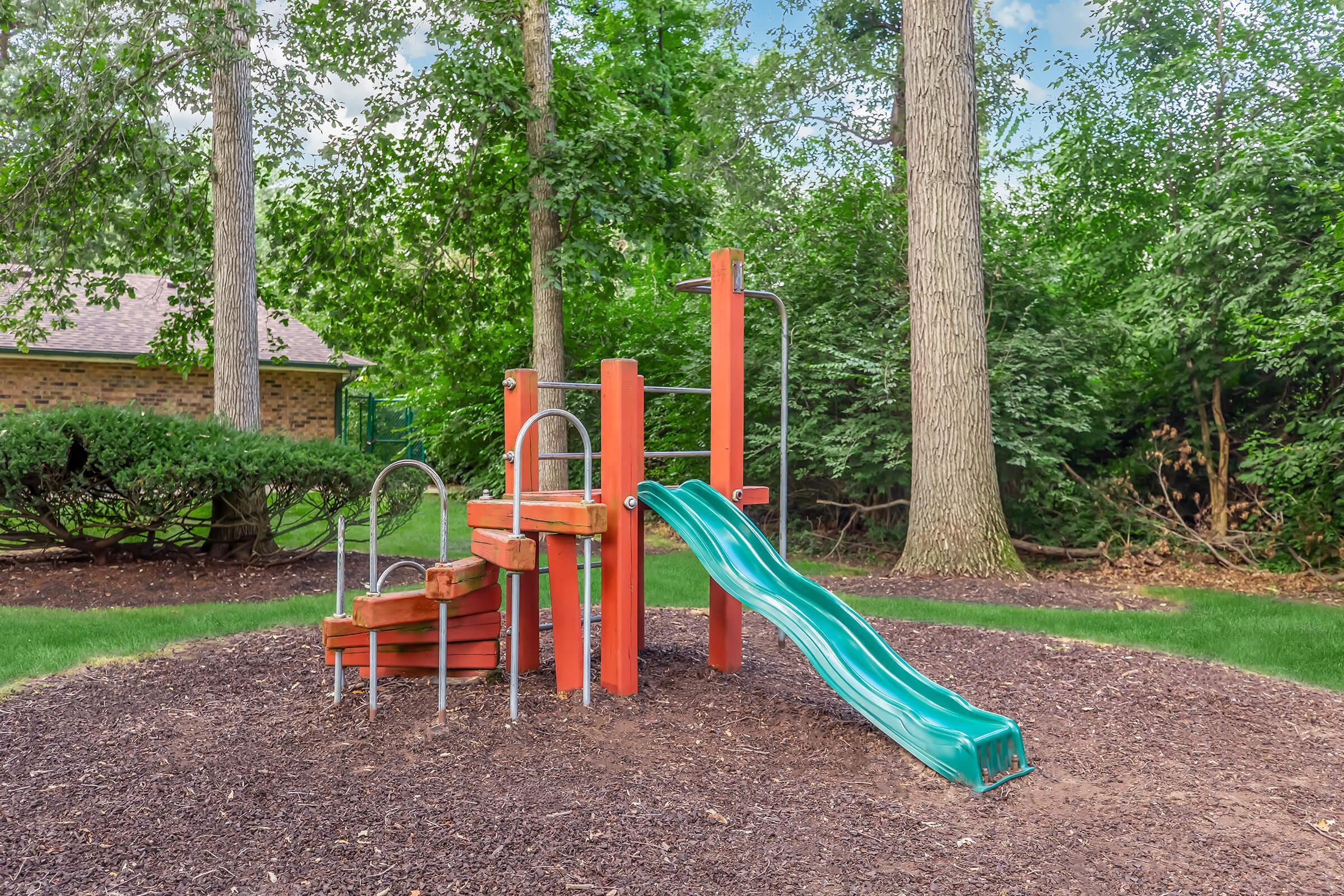 Children's playground equipment featuring a red structure with a green slide, surrounded by mulch and trees. The area appears well-maintained, with a grassy lawn and shrubs in the background, providing a playful outdoor environment.