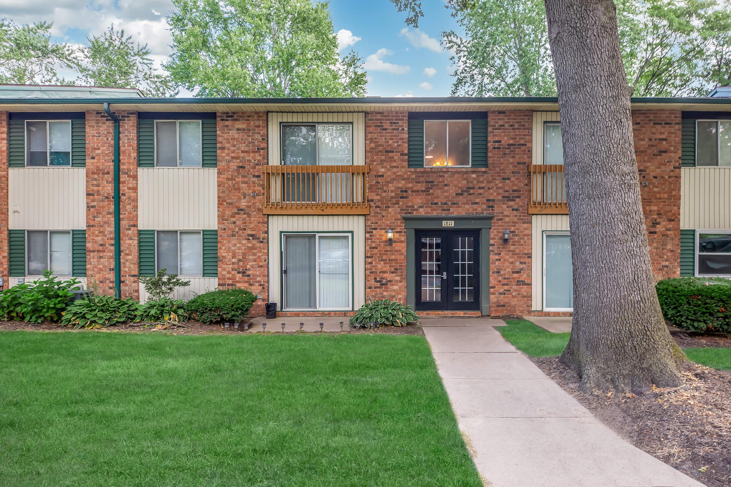 Two-story brick apartment building with green shutters, featuring a central entrance flanked by two balconies. Lush green lawn and neatly trimmed shrubs surround the pathway leading to the front door. Trees provide shade in the foreground against a bright blue sky with clouds.
