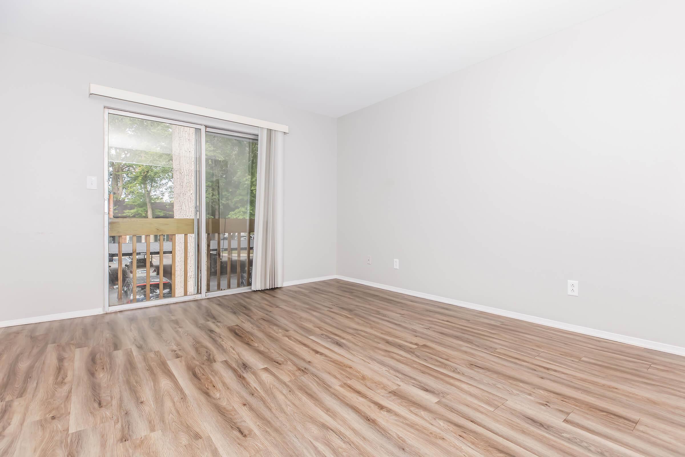 An empty room with light gray walls and laminate flooring. There is a large sliding glass door leading to a small balcony, allowing natural light to fill the space. The room is unfurnished, creating a spacious feel.