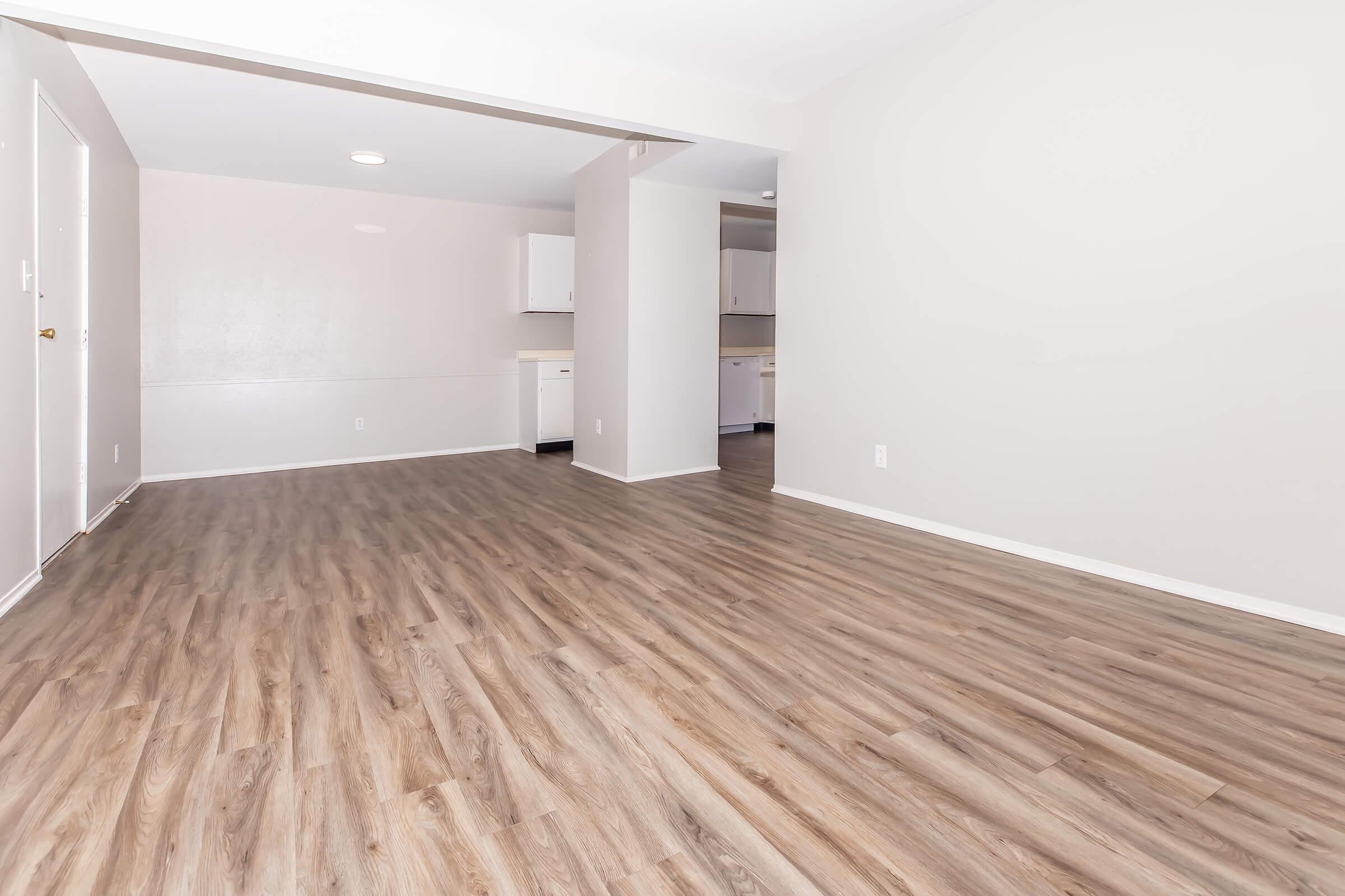 A spacious, empty living area featuring light-colored walls and wooden laminate flooring. The room has natural light coming from a window, and an open layout that leads into a kitchen area with white cabinetry in the background.