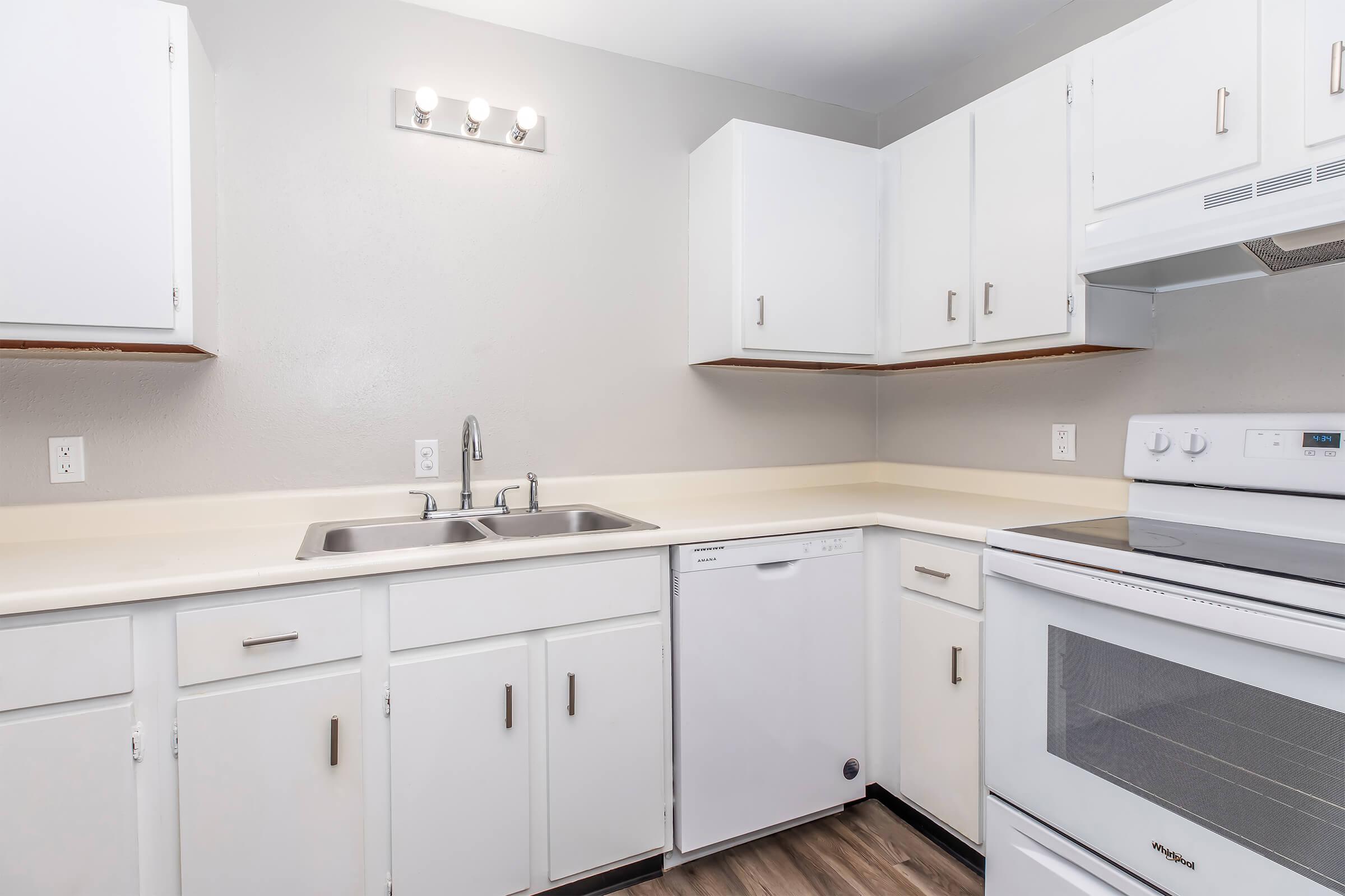 Bright kitchen featuring white cabinets, a double sink, a dishwasher, and a white stove. The countertops are light-colored, and the walls are painted a soft gray. A light fixture is mounted above the sink area, providing ample illumination. The flooring is a dark wood laminate.