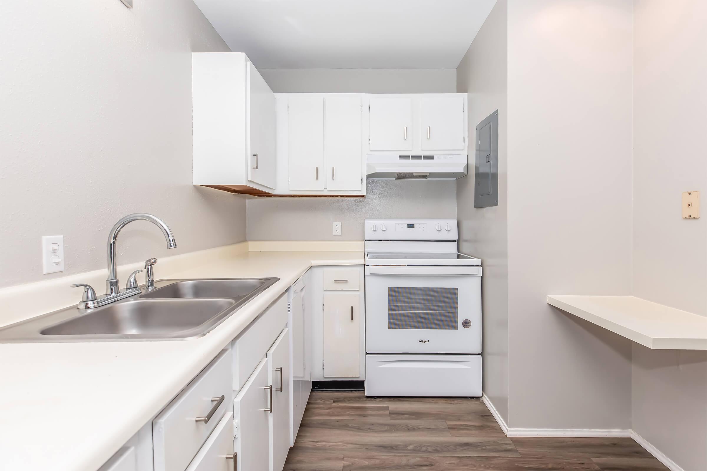 A modern kitchen with white cabinets, a double sink, an electric stove, and a microwave. The countertops are light-colored, and there is a small shelf on the right side. The flooring is a wood-like laminate. The space is well-lit and organized, providing a clean and functional cooking area.