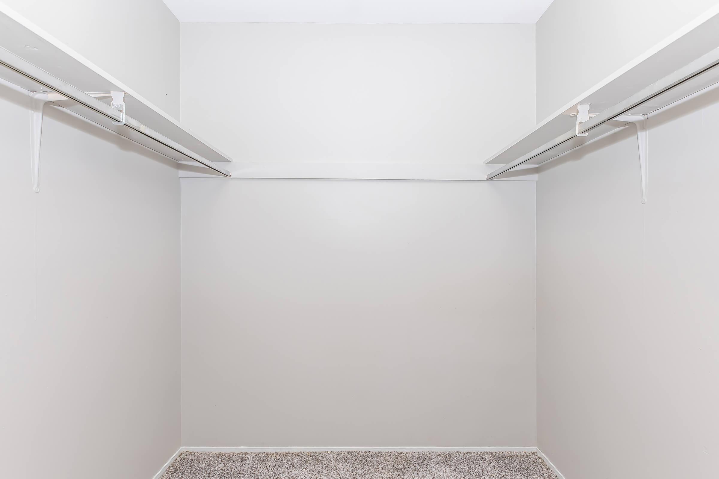 An empty closet with light gray walls and two shelves along the top. The floor is covered with a light-colored carpet. The space is uncluttered and has a minimalistic design, giving a sense of openness.