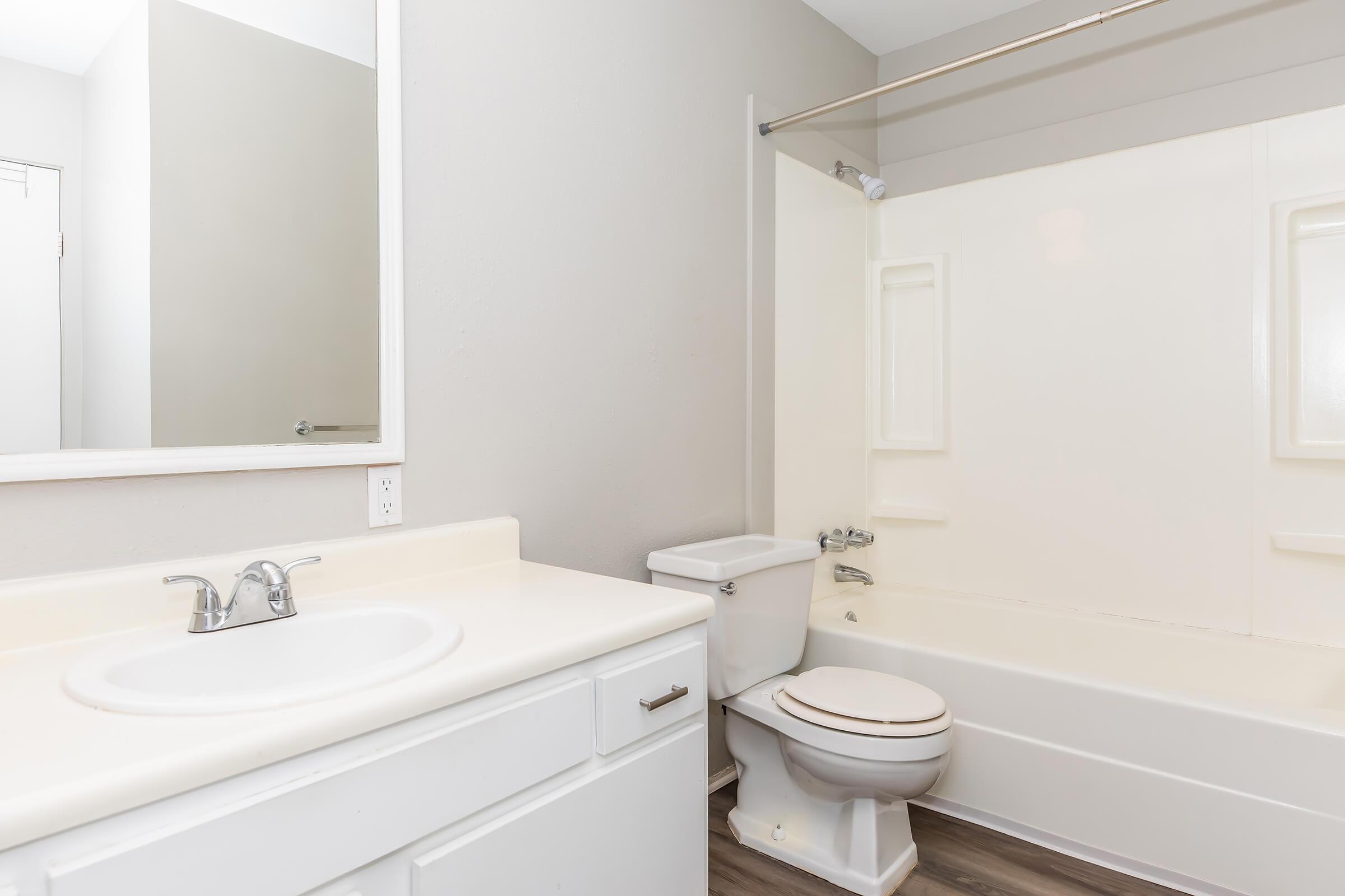 A clean and modern bathroom featuring a white sink with a faucet, a toilet, and a bathtub with a shower. The walls are painted in a light gray color, and there is a large mirror above the sink. The floor has a wood-like finish, contributing to the contemporary design.