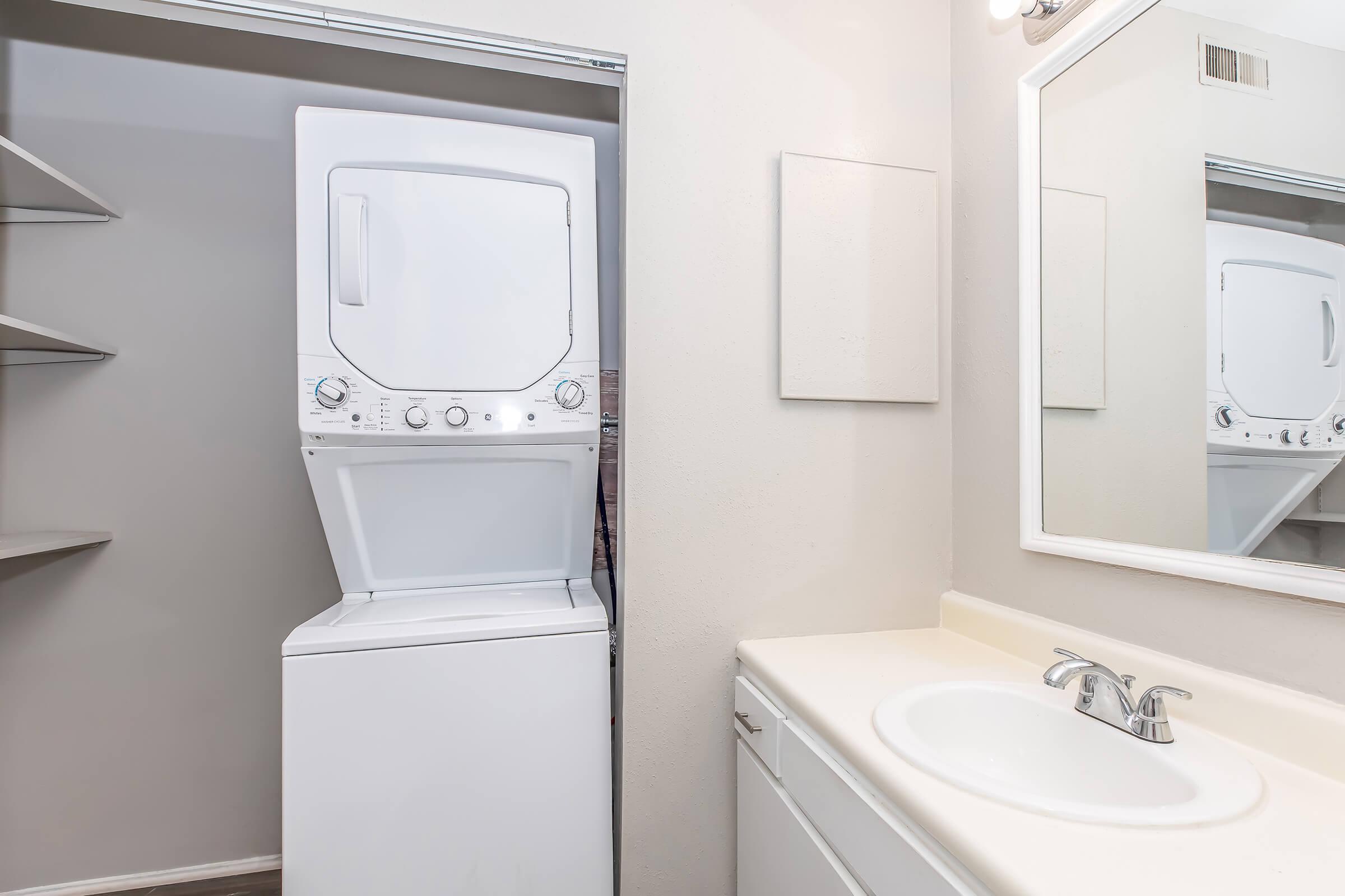 A laundry area featuring a stacked washer and dryer unit next to a white bathroom vanity with a sink. The walls are painted light gray, and there is a mirror above the sink. Shelving is visible to the left, contributing to the overall functional design of the space.