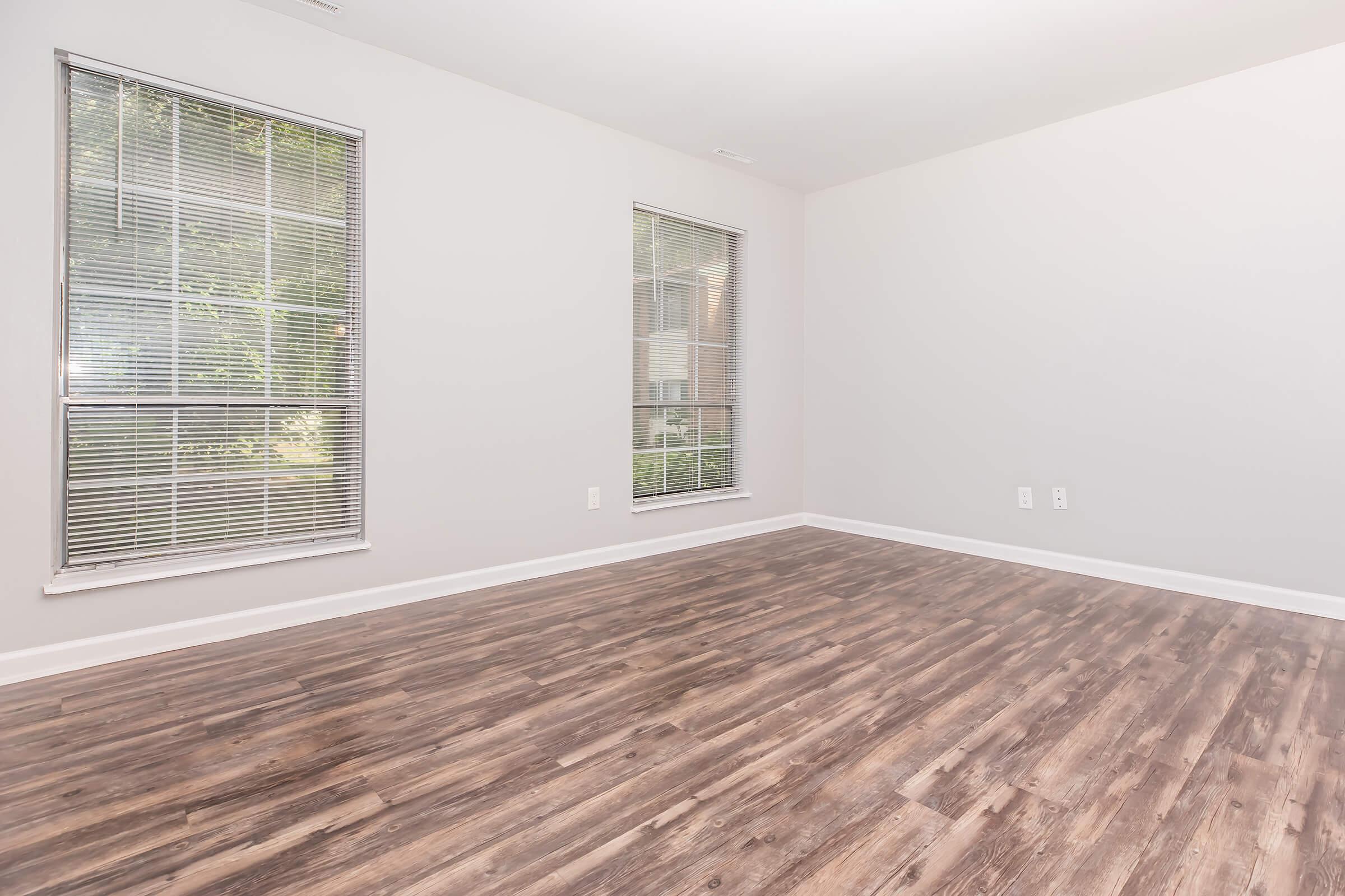 Empty room with light gray walls and two windows with blinds allowing natural light. The floor features wooden laminate flooring, creating a warm and inviting atmosphere. The space is uncluttered, giving it a spacious and minimalist feel.