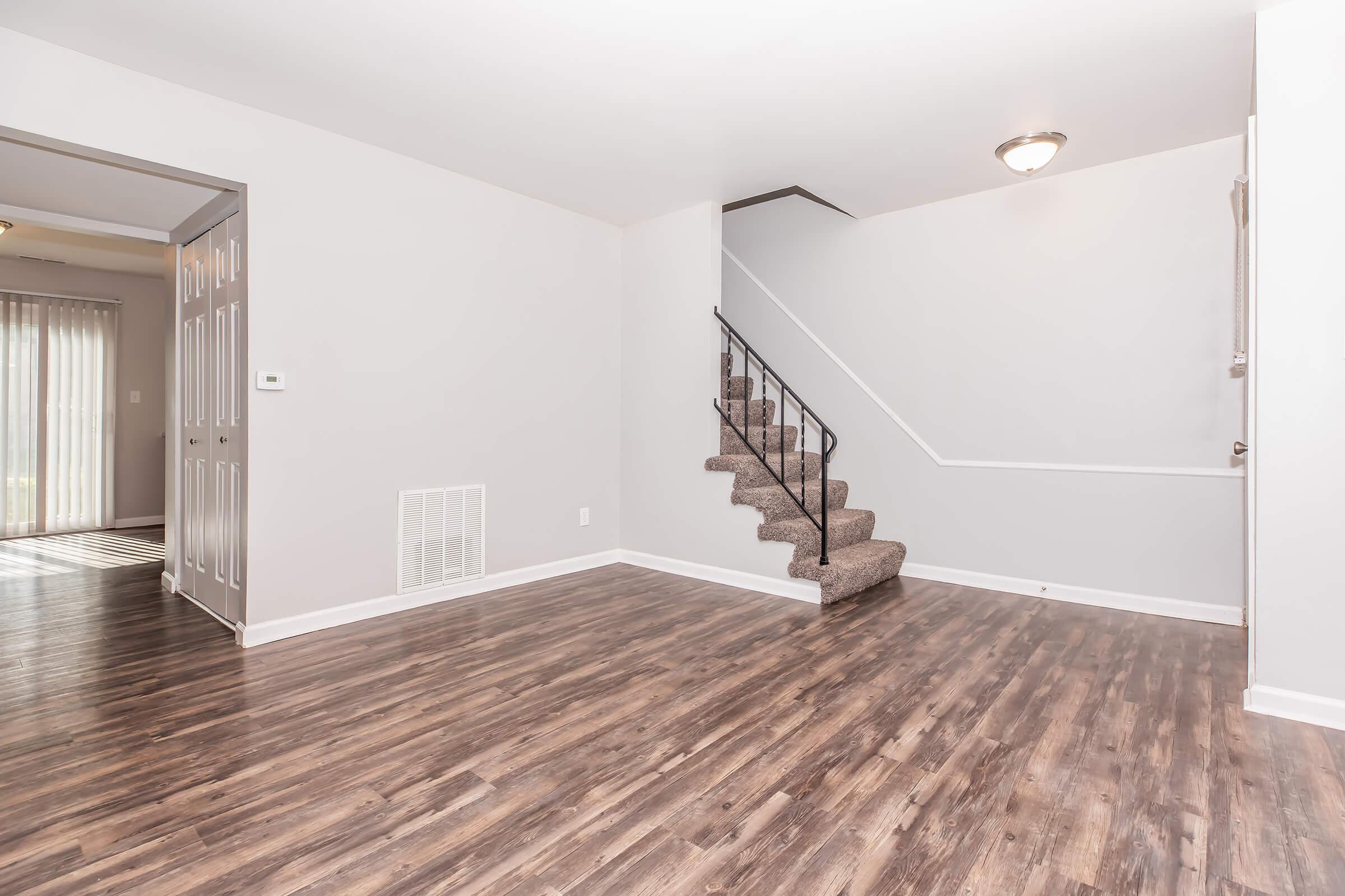 A spacious, well-lit interior room featuring hardwood-style flooring. On the left, there's a doorway leading to another area with sliding glass doors. A staircase with a black railing ascends along the right wall, and the walls are painted in a light, neutral color, contributing to a fresh and modern look.