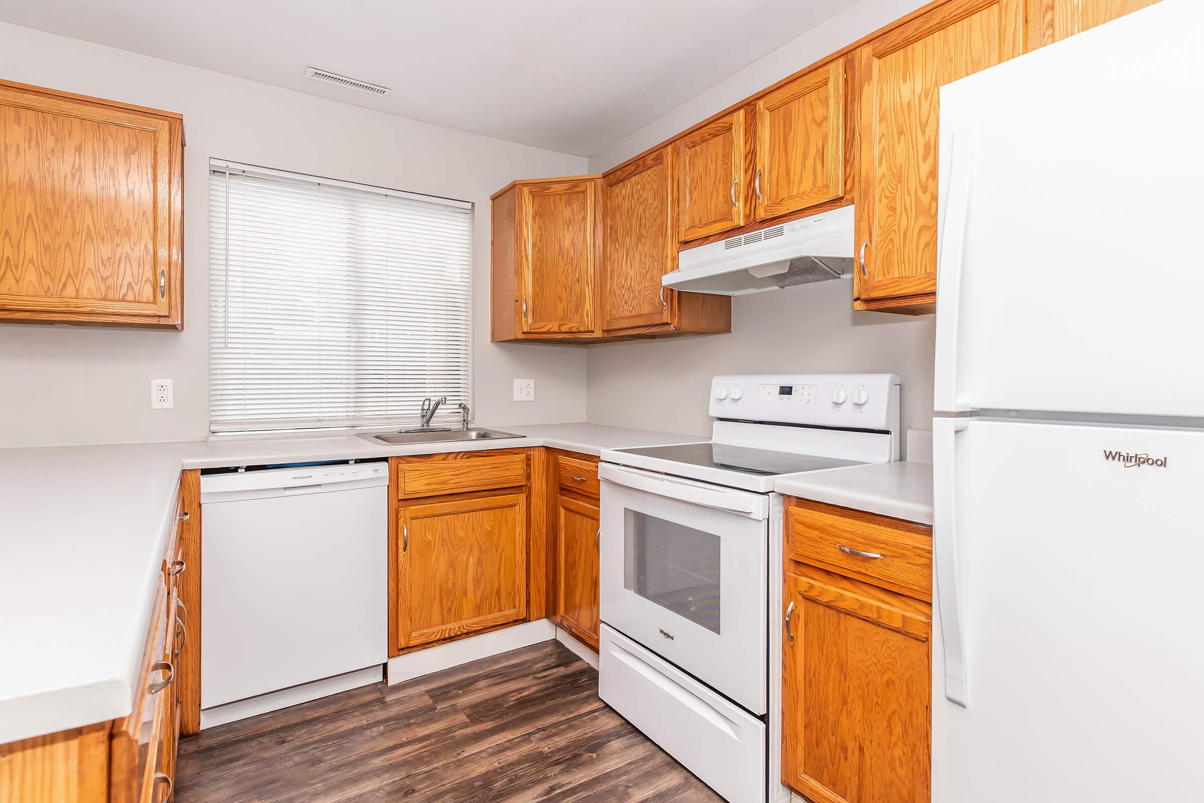 A well-lit kitchen featuring wooden cabinetry, a white refrigerator, a dishwasher, and an electric stove. The countertop is gray, and the window has blinds. The flooring is a dark wood laminate, creating a warm and inviting atmosphere.