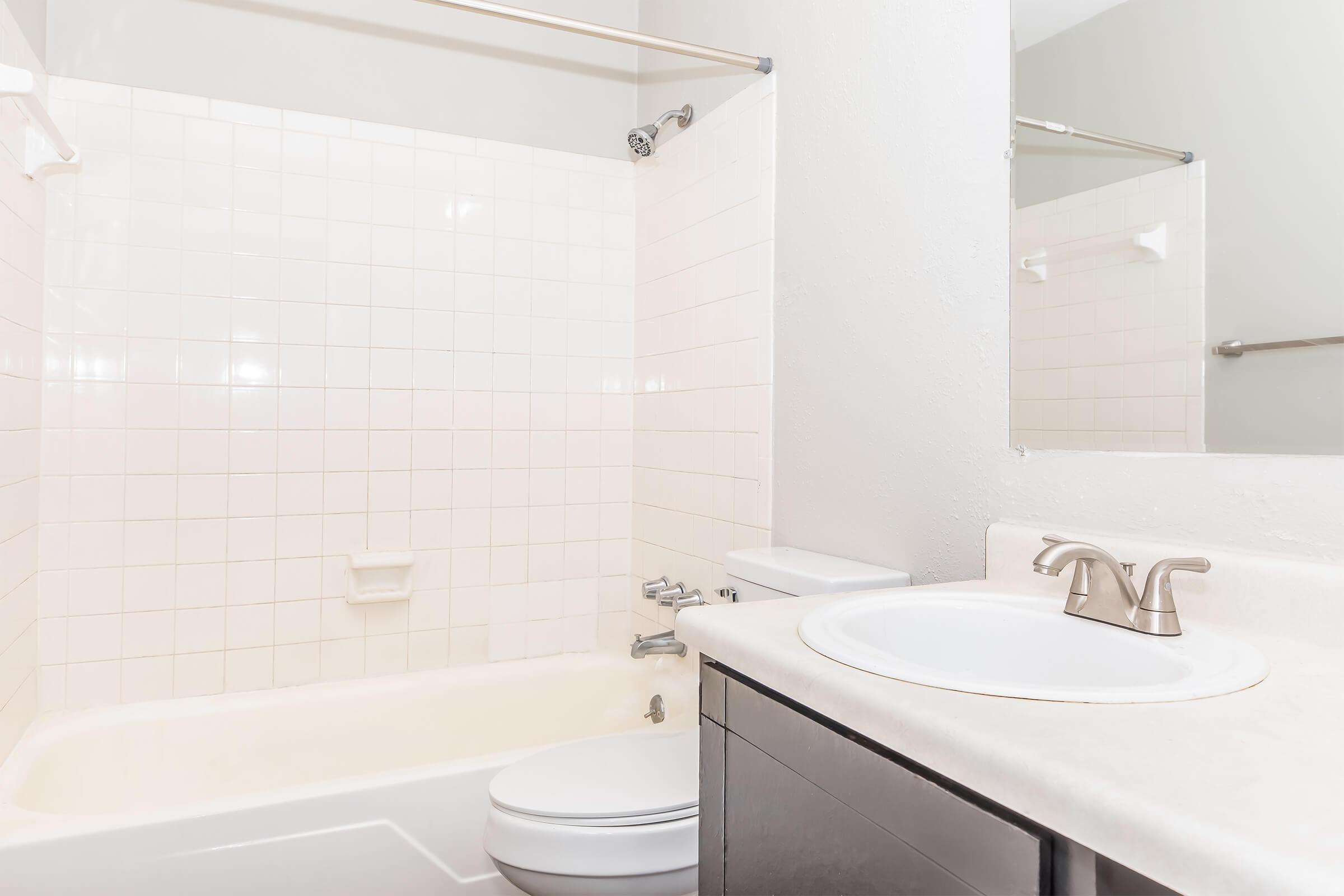 A clean, well-lit bathroom featuring a white bathtub with a shower curtain, a sink with a modern faucet, and gray walls. The space has white tiled walls and minimalistic design elements, providing a fresh and tidy appearance. A mirror is mounted above the sink, reflecting the bathroom setting.