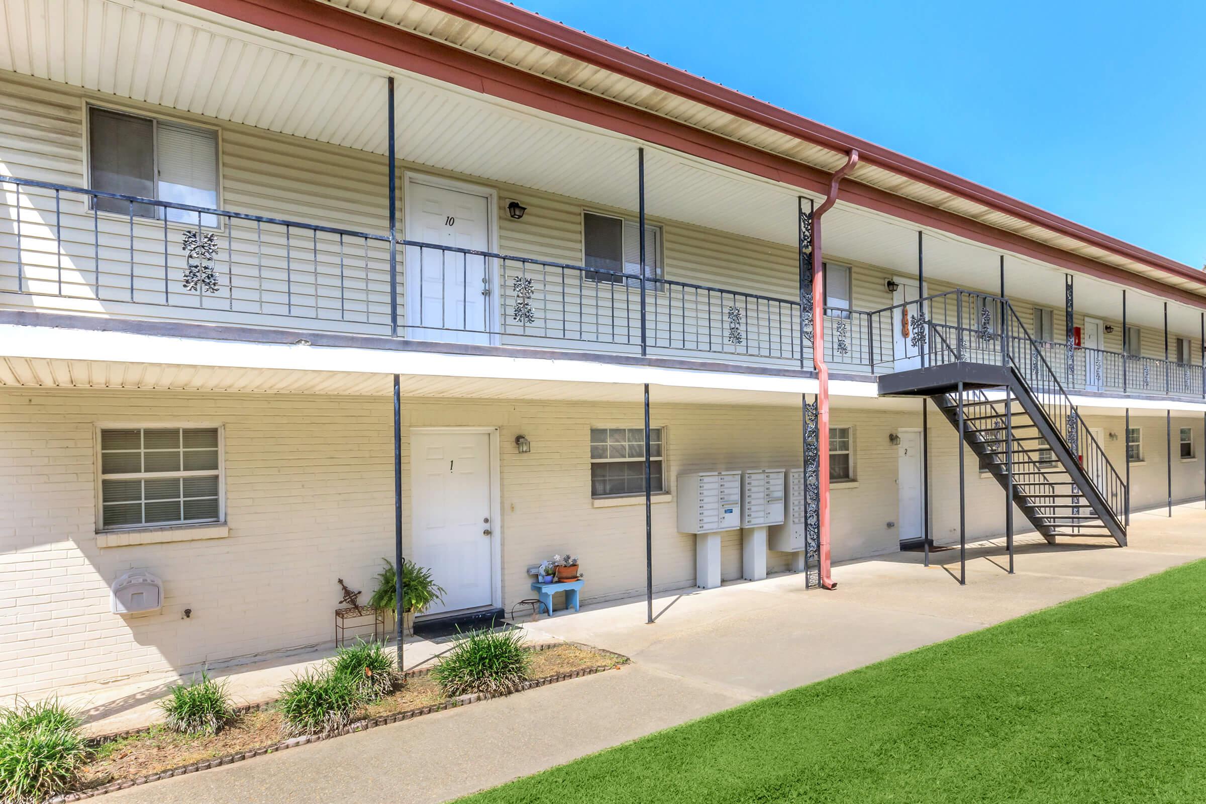 A view of a two-story apartment complex with a porch, featuring several doors and a staircase leading to the second floor. The exterior walls are light-colored brick, with a neatly maintained lawn and shrubs in front. Bright blue sky in the background enhances the scene.