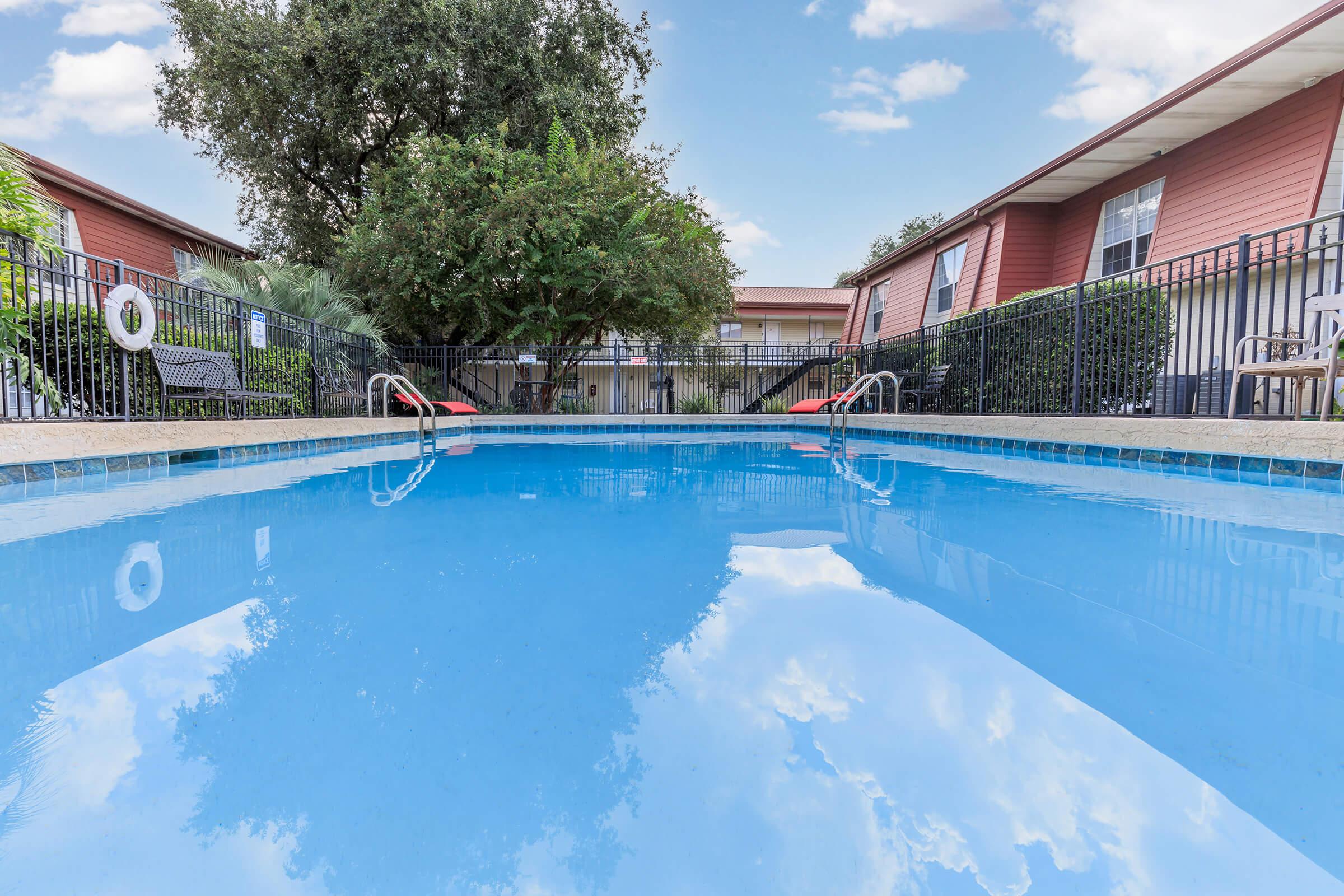 A clear blue swimming pool with a smooth surface reflecting the sky, surrounded by a black fence. In the background, there are trees and buildings with red exteriors. The scene is calm and inviting, perfect for relaxation. A lifebuoy is visible on the fence nearby.