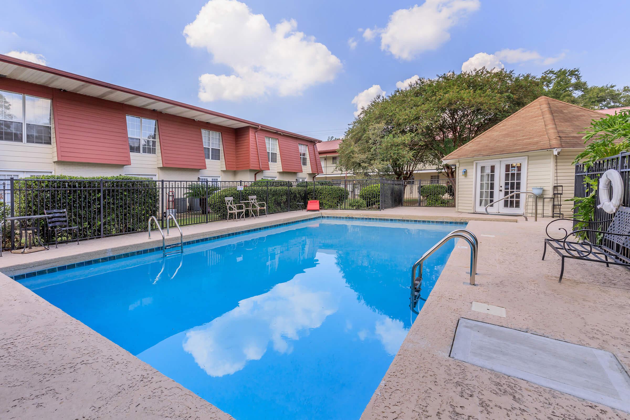 A clear blue swimming pool surrounded by a fenced area, with lounge chairs on one side. In the background, there are two-story residential buildings with red accents. A small gazebo or clubhouse is visible at one end of the pool area, set against a backdrop of trees and a partly cloudy sky.