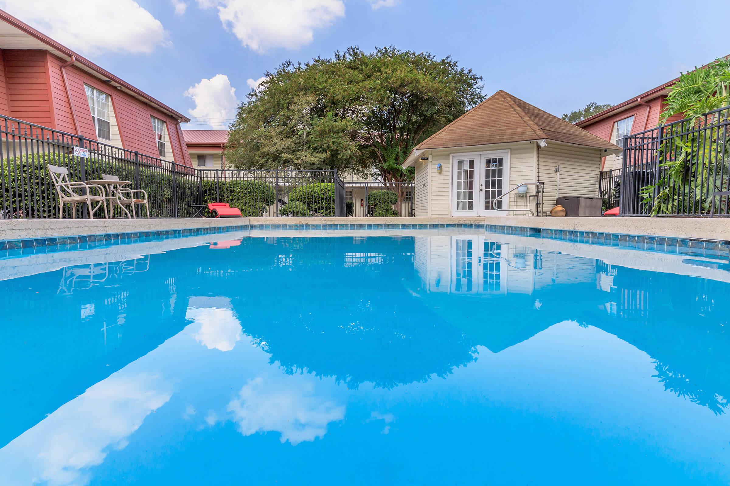 Clear blue swimming pool reflecting the sky, surrounded by a landscaped area with trees and a gazebo. In the background, red and beige buildings with outdoor seating can be seen. The tranquil scene suggests a relaxing atmosphere perfect for leisure and outdoor enjoyment.