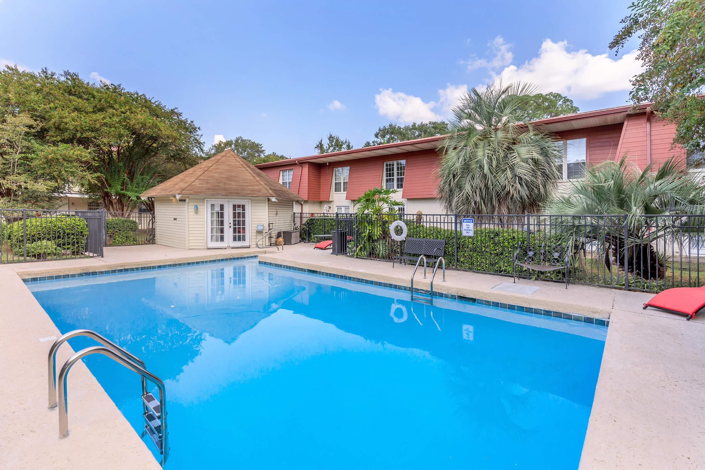 A tranquil swimming pool surrounded by palm trees and a well-kept garden, featuring a small gazebo nearby. The pool has a ladder for entry and patio chairs along the edge, with a clear blue sky above.
