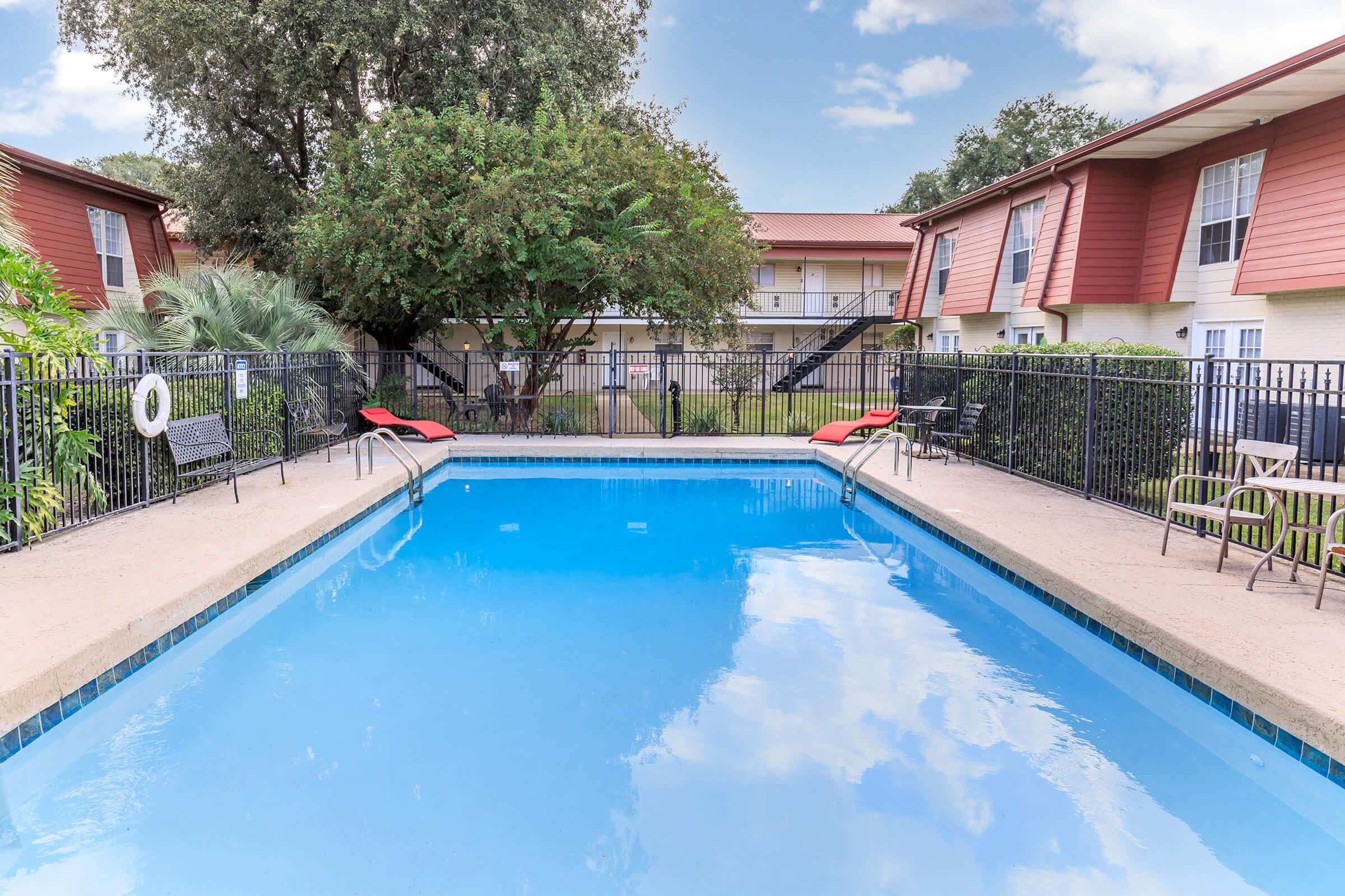 A clear blue swimming pool surrounded by a black metal fence, with lounge chairs on either side. Lush green trees and foliage are visible in the background, along with a two-story red and white building. The sky is partly cloudy, creating a serene atmosphere.