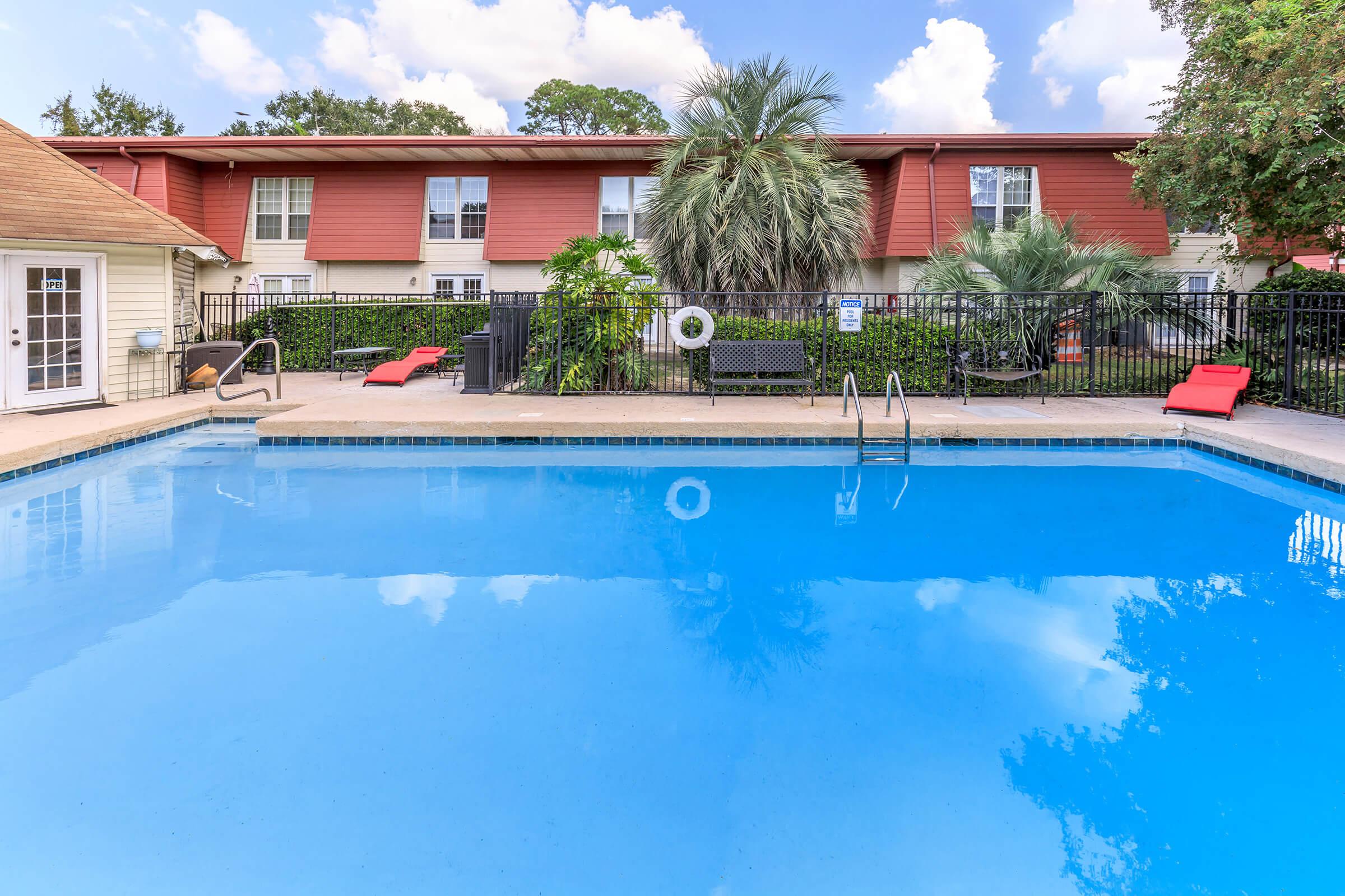A tranquil swimming pool surrounded by a black fence, with two red lounge chairs positioned near the edge. There are vibrant palm trees and green shrubs in the background, and a two-story building with a red exterior is visible in the distance under a partly cloudy blue sky.