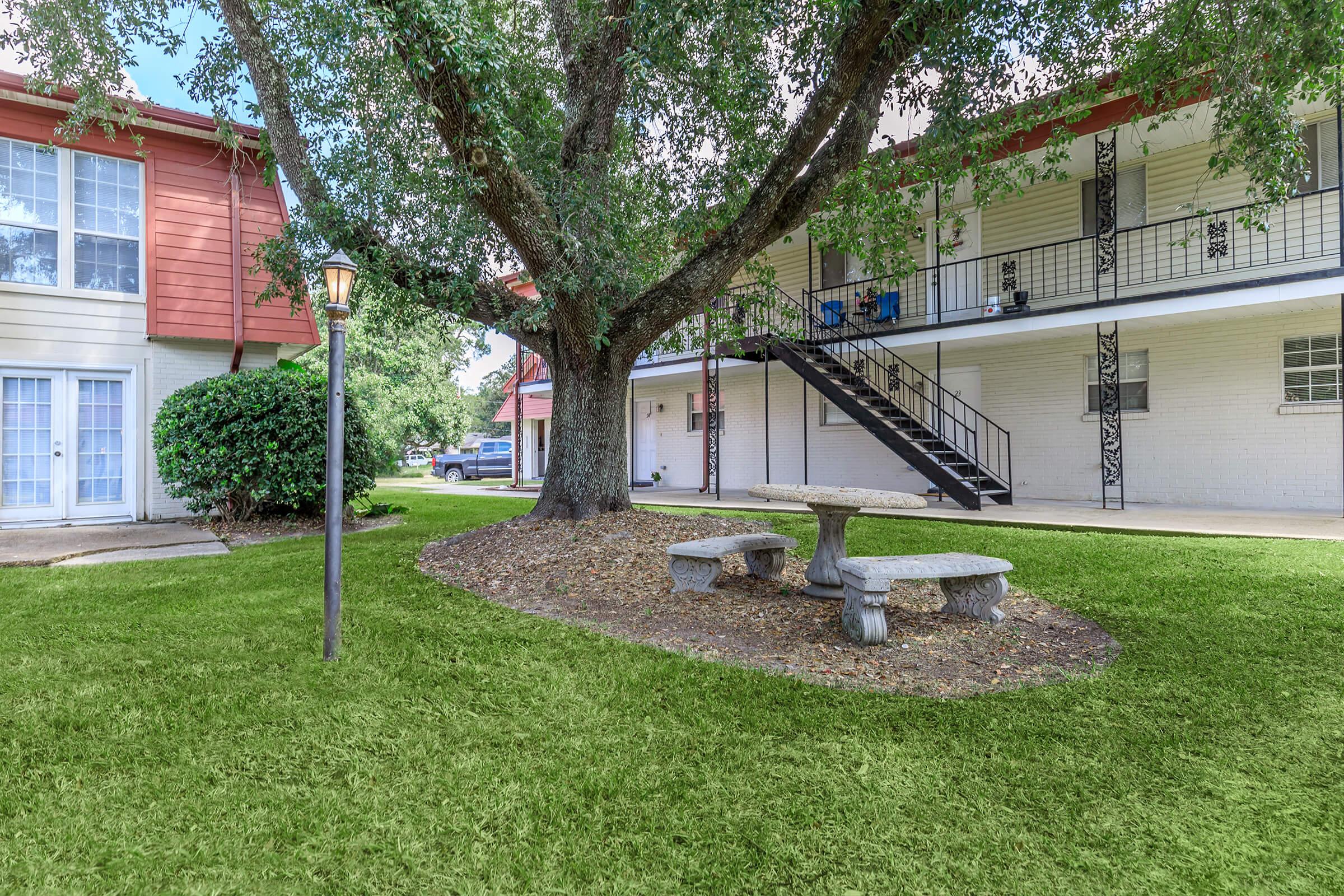 A landscaped area featuring a large tree and a stone table with benches underneath. In the background, there are two buildings, one with red siding and another with white, alongside a black metal staircase. The scene is set in a well-maintained green lawn.