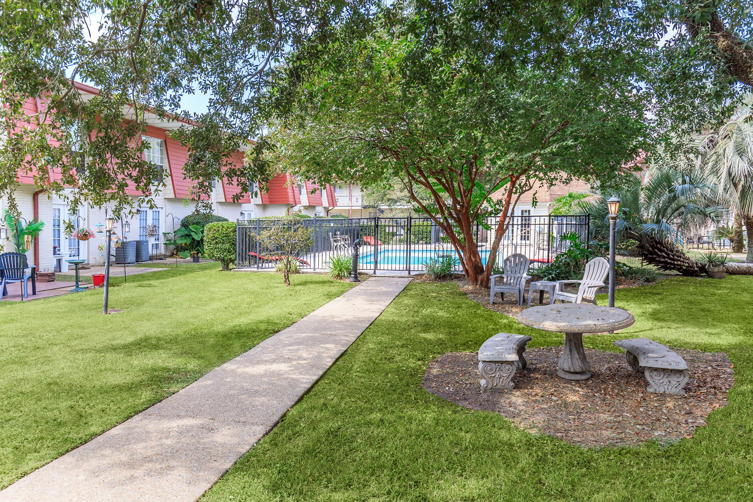A landscaped garden area featuring a concrete pathway leading to a gated swimming pool. There are several trees for shade, a stone table surrounded by chairs, and a green lawn with decorative plants. In the background, there are buildings with red accents.