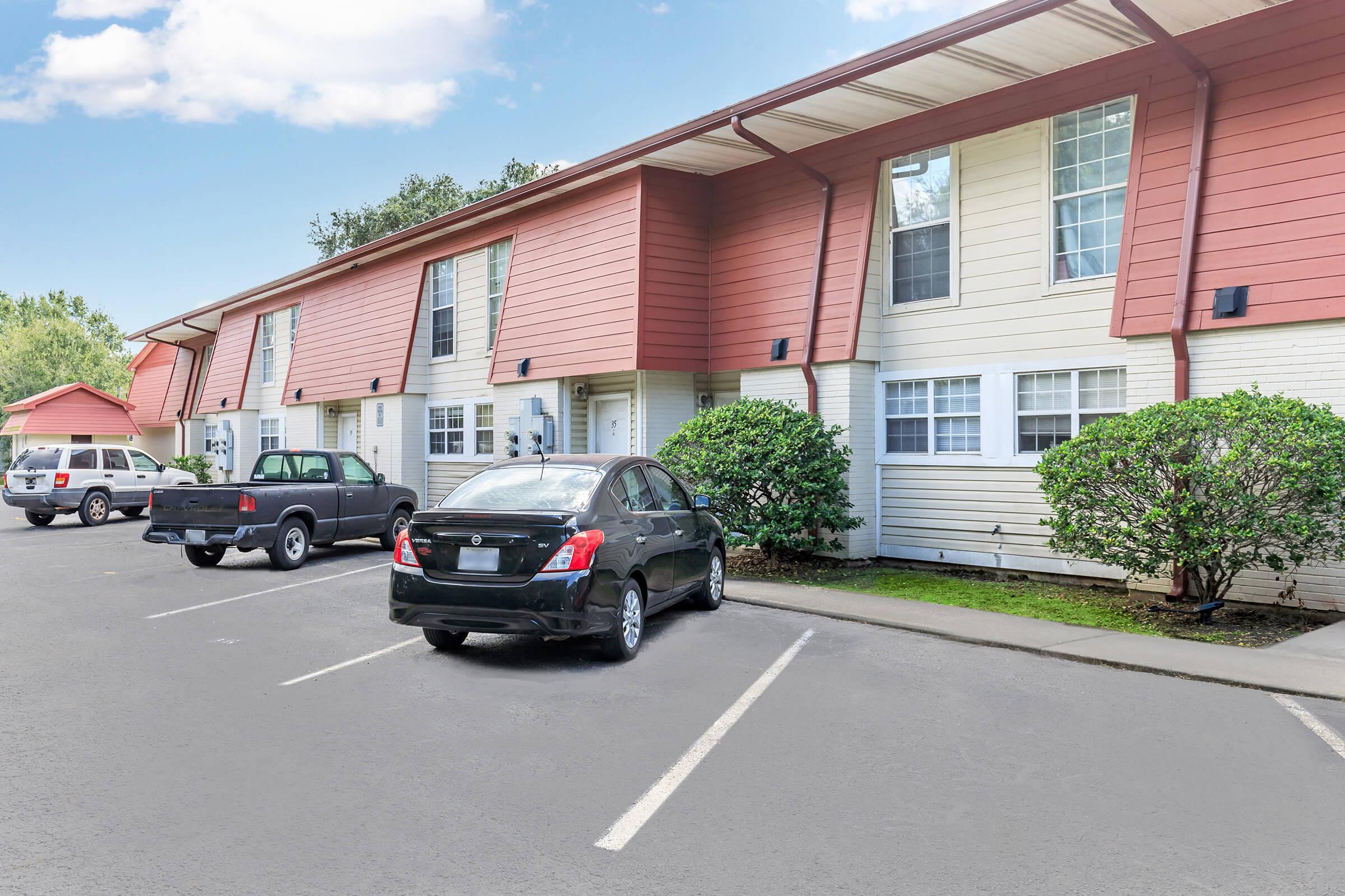 A row of two-story apartment buildings with red and beige siding. Cars are parked in the foreground, including a black sedan and a pickup truck. The area features small shrubs and trees, with blue sky and fluffy clouds visible above. The scene depicts a quiet residential environment.