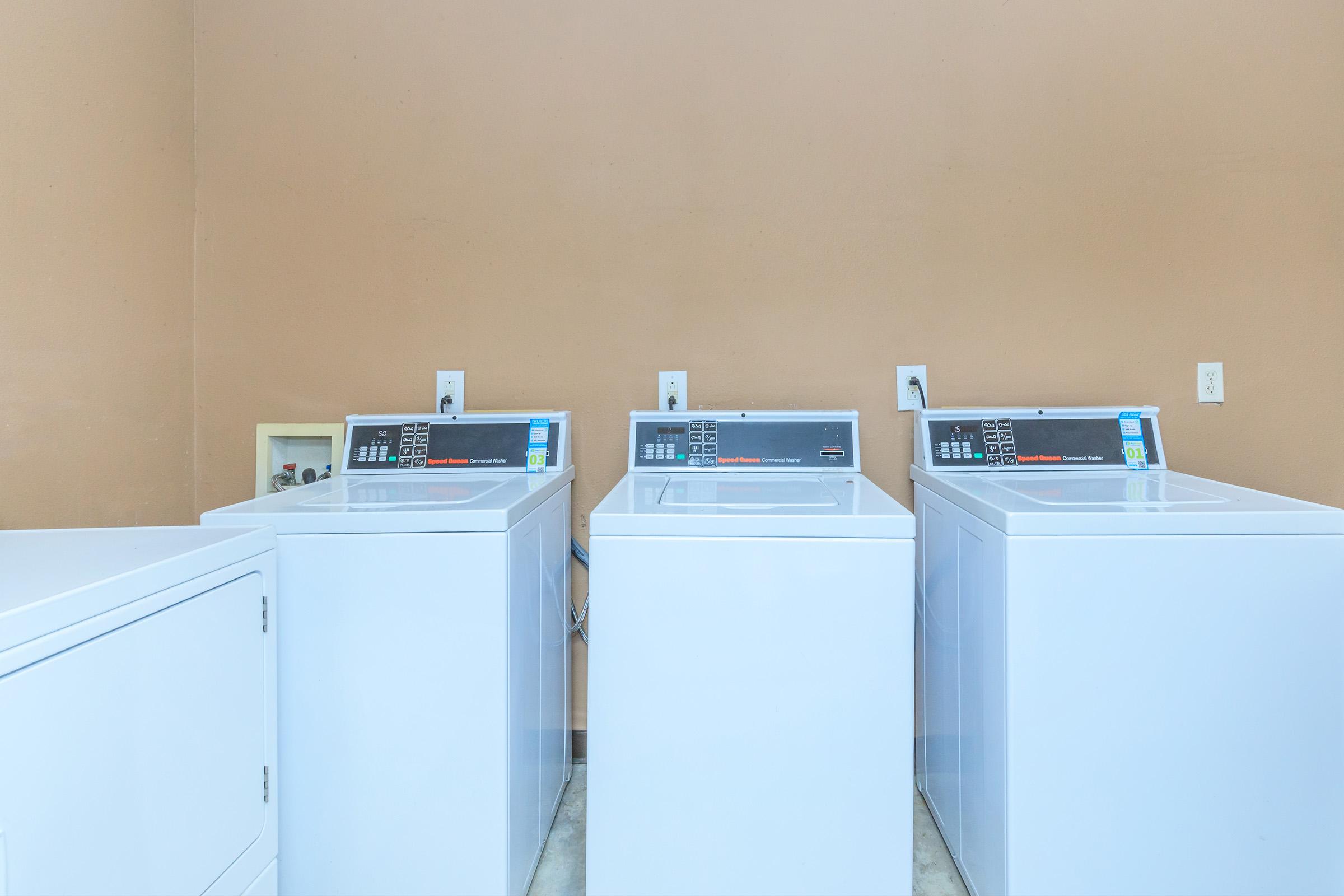 Three white washing machines lined up against a light brown wall in a laundry room. Each machine has digital displays and control panels on top, with a utility sink visible on the left. The floor is tiled, and electrical outlets are present on the wall. Overall, the space is clean and organized.