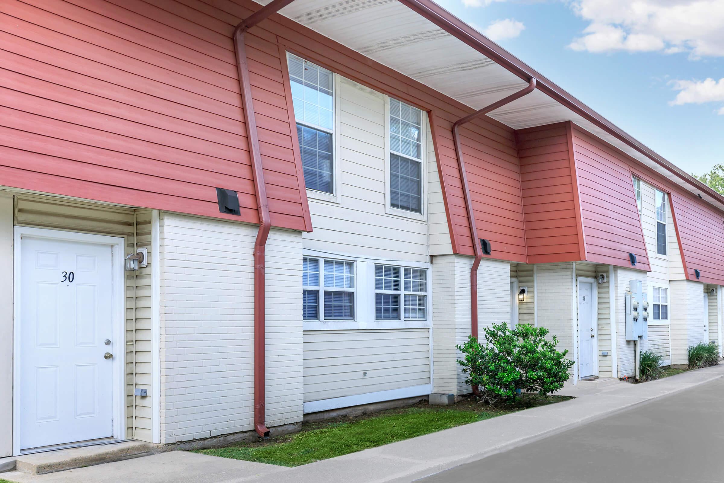 Row of townhouses with a mix of light-colored siding and red roofs. Each unit has windows and a front door, and there's a small shrubbery along the pathway. The setting appears well-maintained with a clear sky in the background.