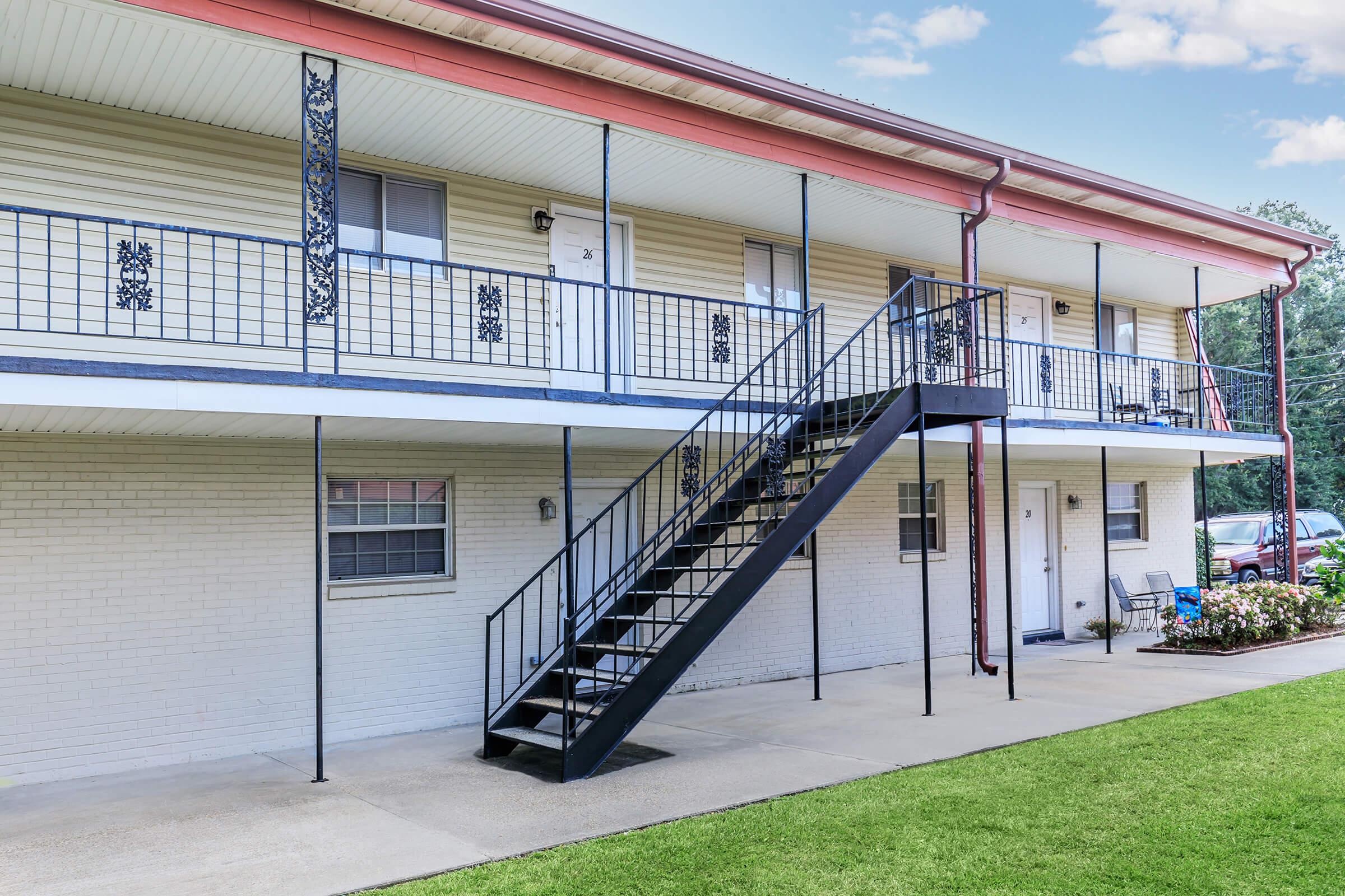 Exterior view of a two-story apartment building featuring a black metal staircase leading to the second floor. The building has a light-colored exterior with white brick, balconies with decorative railings, and a well-maintained lawn in front. Several doors and windows are visible on each floor.