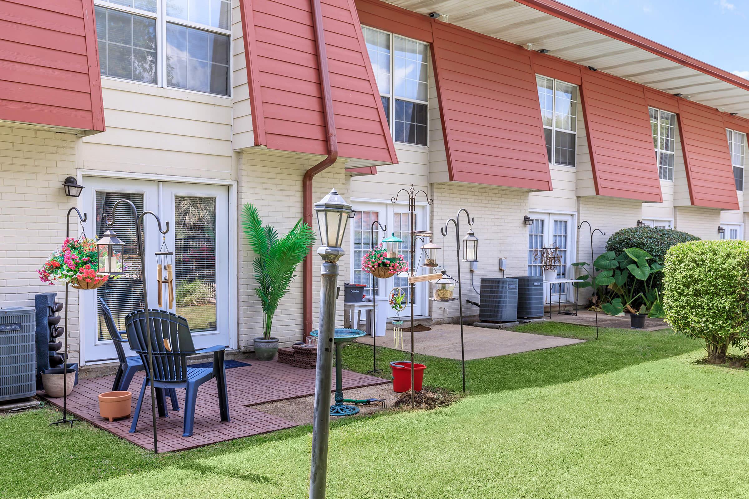 A cozy outdoor patio area with a few potted plants and a black chair. Several bird feeders and hanging flower baskets adorn the space, with a well-maintained lawn in the foreground. The building features a distinct red awning and large windows, creating a welcoming atmosphere.