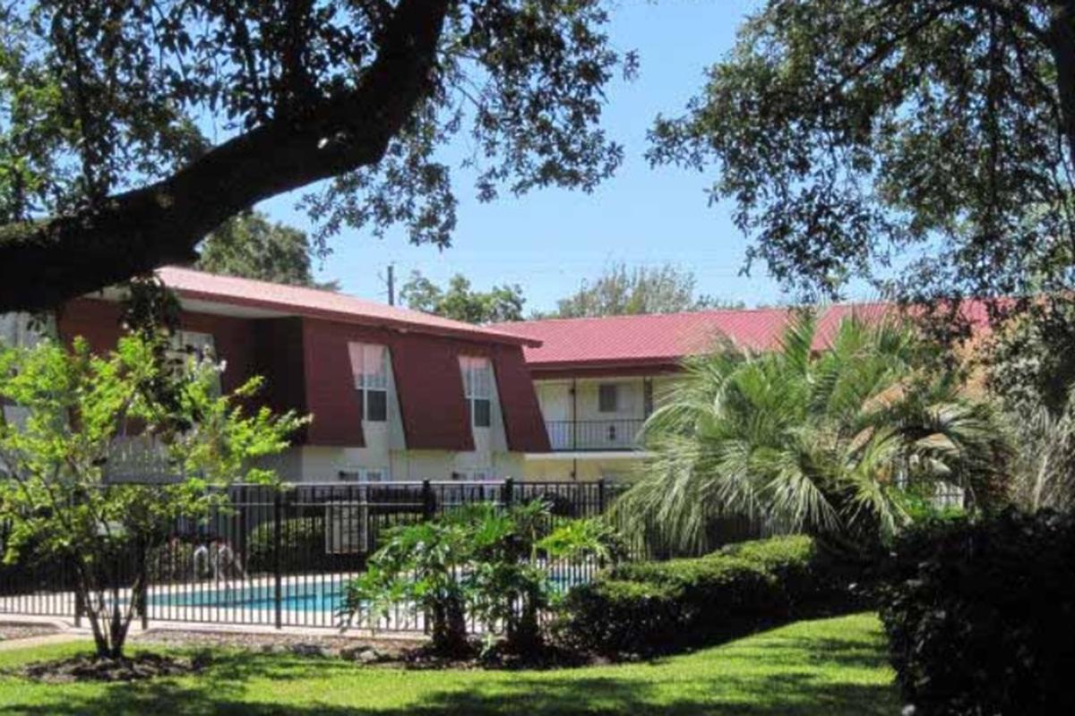 A view of a residential building with a reddish-brown exterior and a red metal roof, surrounded by lush greenery, palm trees, and a swimming pool enclosed by a black fence. The scene is bright and sunny, with clear blue skies and well-maintained landscaping.