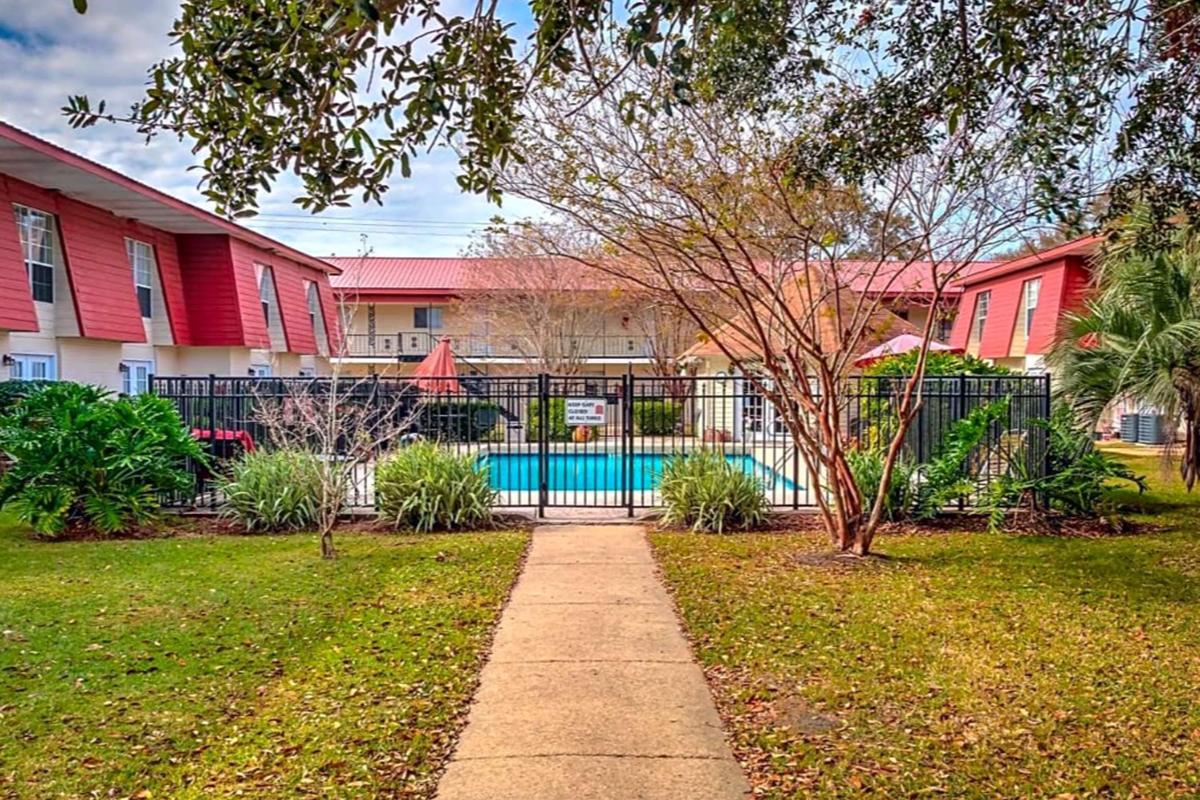 A clear view of a swimming pool surrounded by a black metal fence, with green grass and shrubs in the foreground. The area is framed by two red apartment buildings in the background, and there are a few lounge chairs and shaded areas with umbrellas near the pool. The scenery is peaceful and inviting.