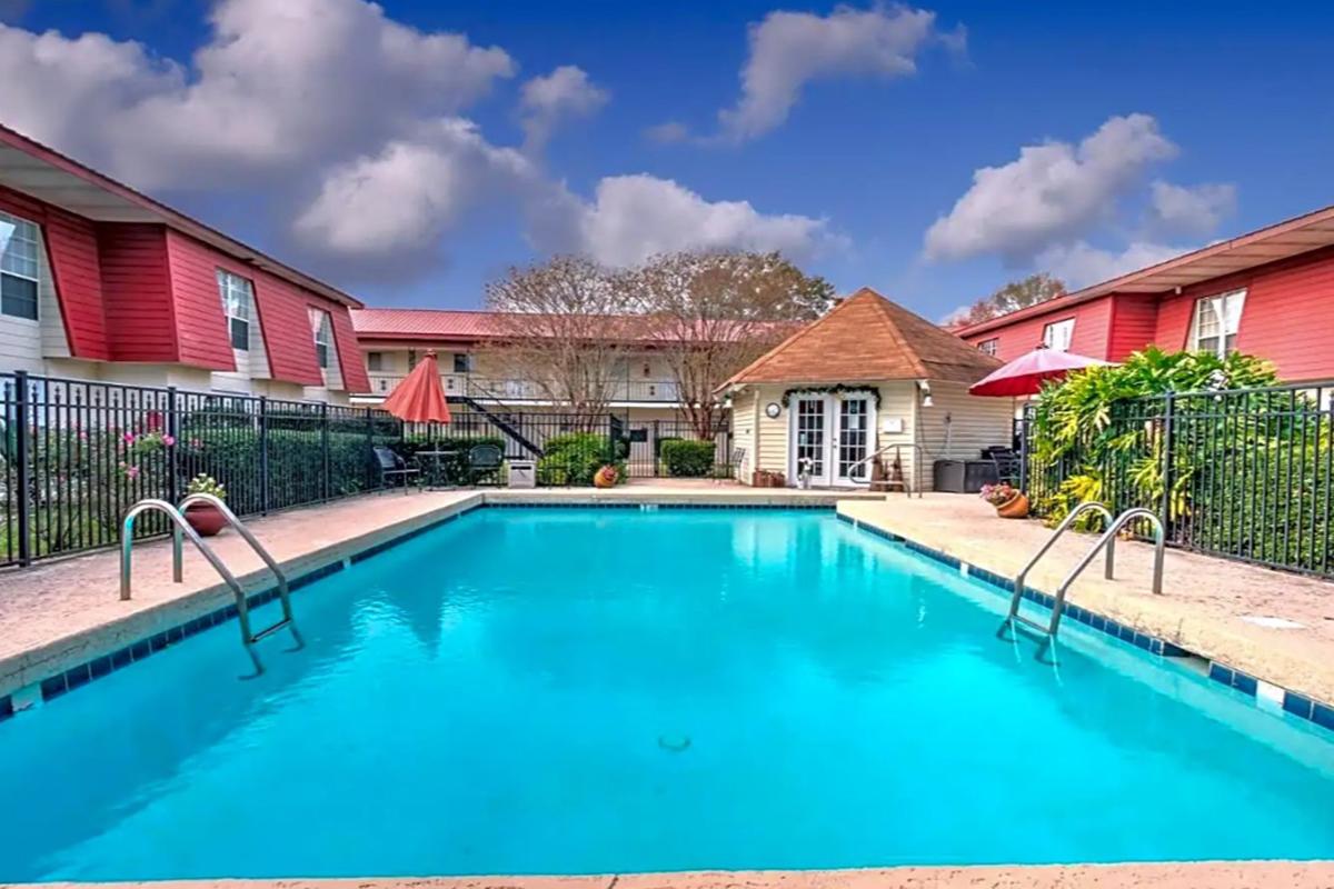 A vibrant swimming pool surrounded by lush greenery and landscaped areas. The pool features two steps leading in, with a wooden gazebo nearby. Red and beige buildings border the pool area, and colorful umbrellas provide shade. A clear blue sky dotted with clouds completes the serene atmosphere.