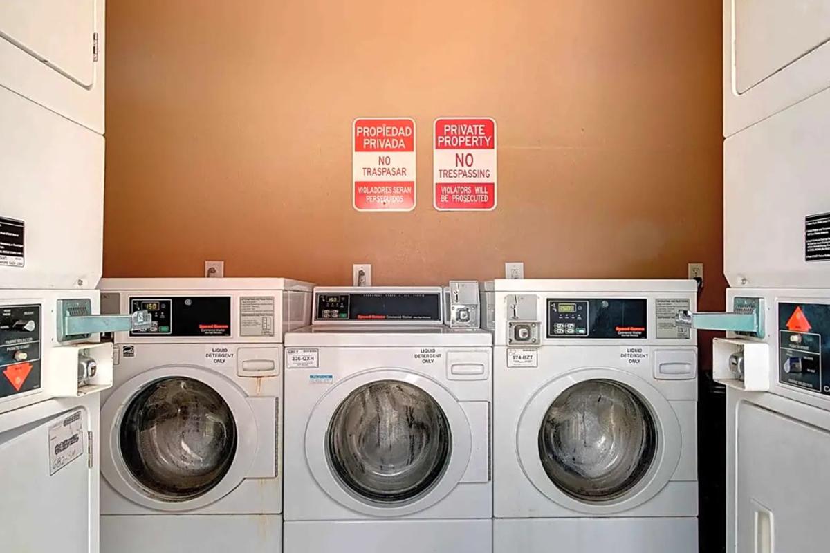 A laundry room featuring three stacked washing machines and dryers. The machines are white and metallic, with visible controls. On the wall above, there are signs in English and Spanish stating "Private Property" and "No Trespassing." The overall atmosphere is utilitarian and tidy.