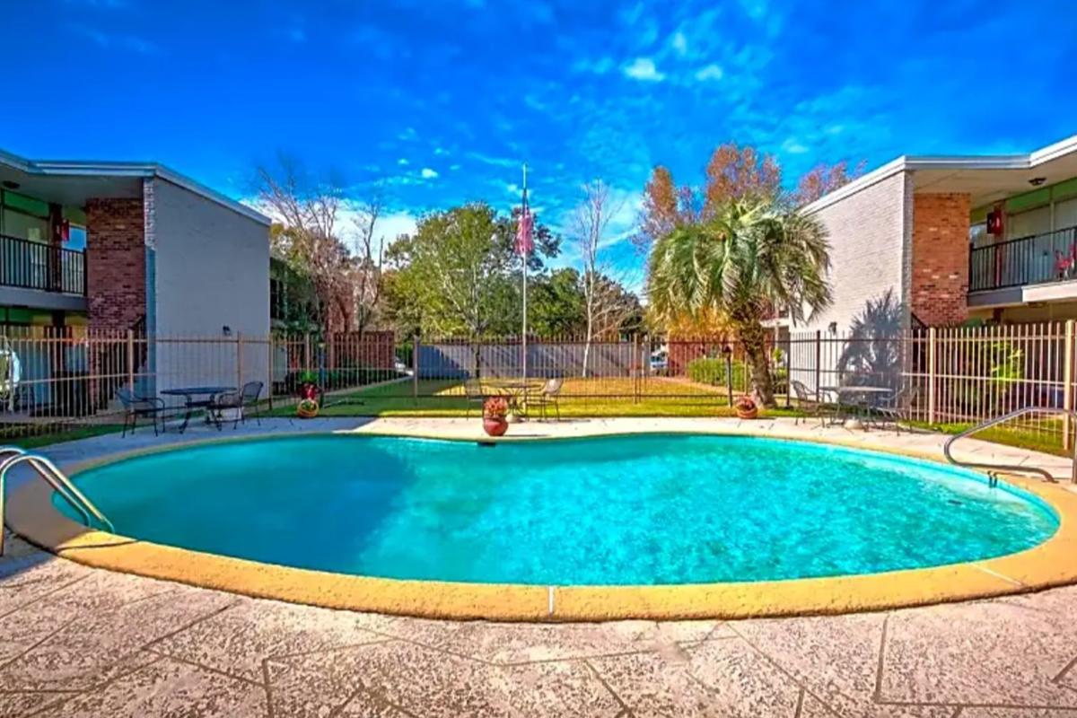A spacious swimming pool surrounded by a patio and greenery, situated between two apartment buildings. The water is clear and inviting, with a bright blue sky and scattered clouds above. Palm trees and well-maintained landscaping enhance the serene atmosphere.